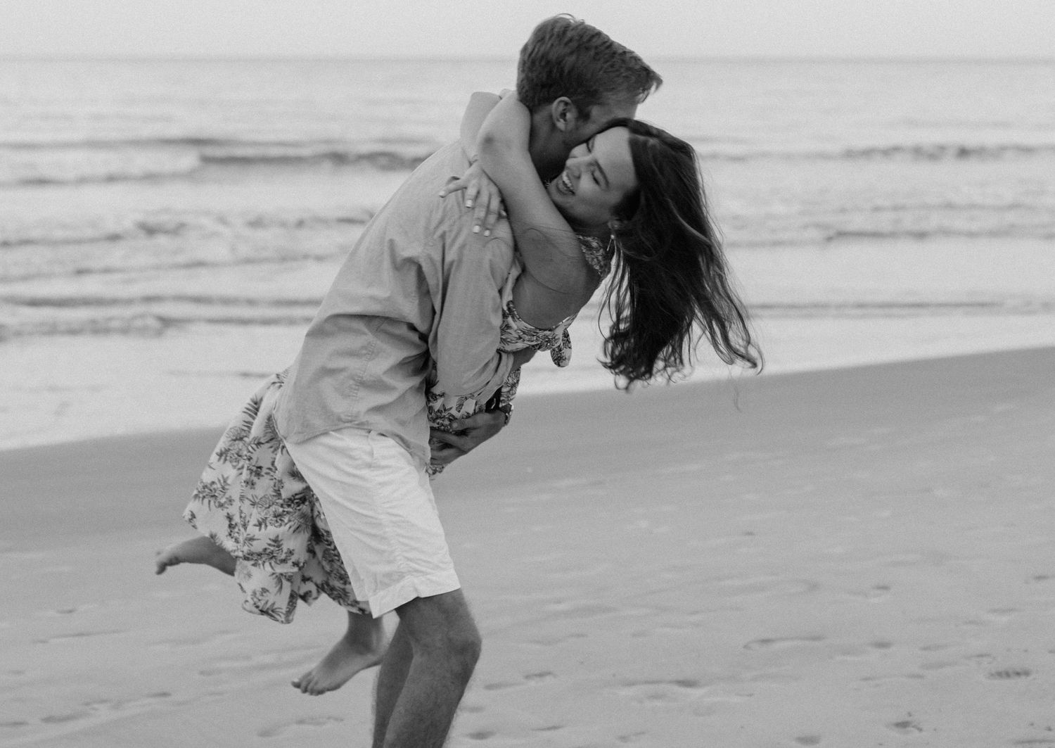 A romantic couple embraces and kisses on the beach in a black and white photograph.