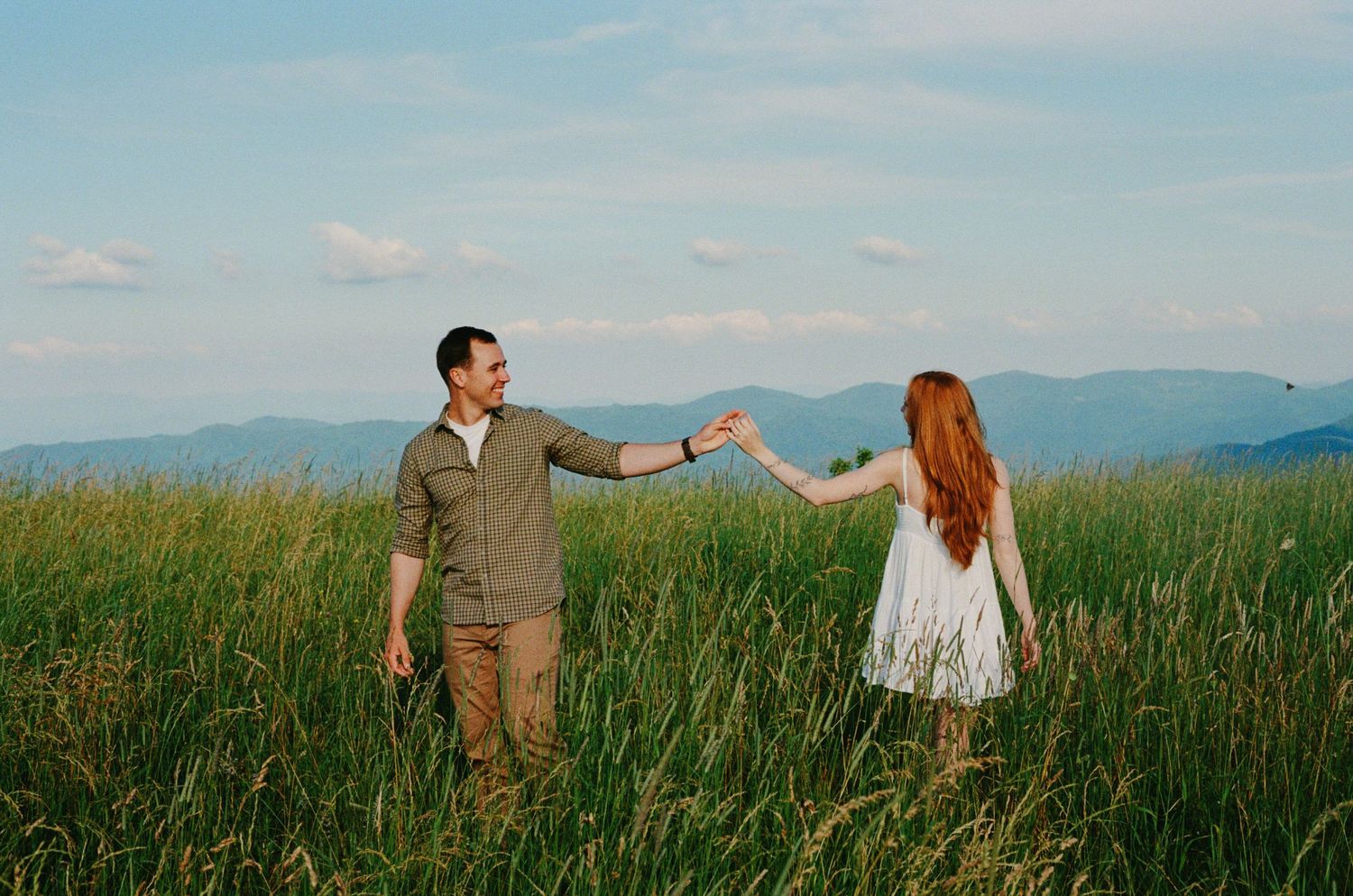 Two people holding hands in a scenic field with mountains and blue sky in the background.