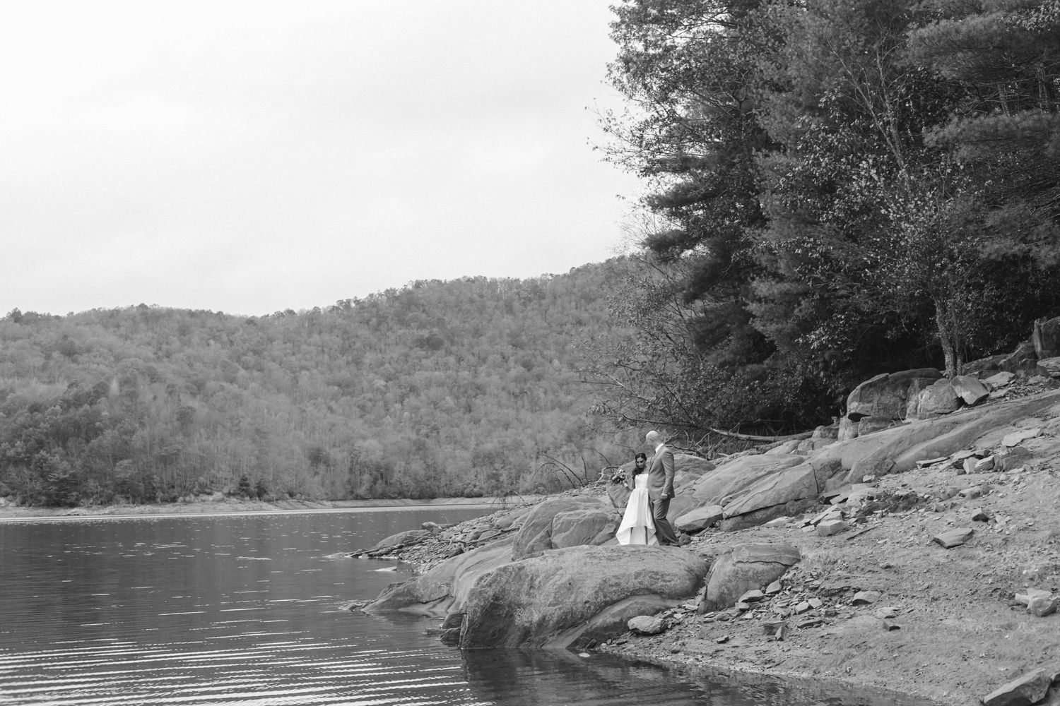 A couple stands on a rocky lakeshore surrounded by wooded hills in a serene black and white landscape.