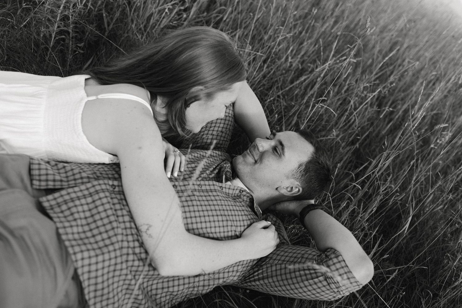 Romantic black and white photo of a couple lying in tall grass, tenderly embracing in intimate moment.