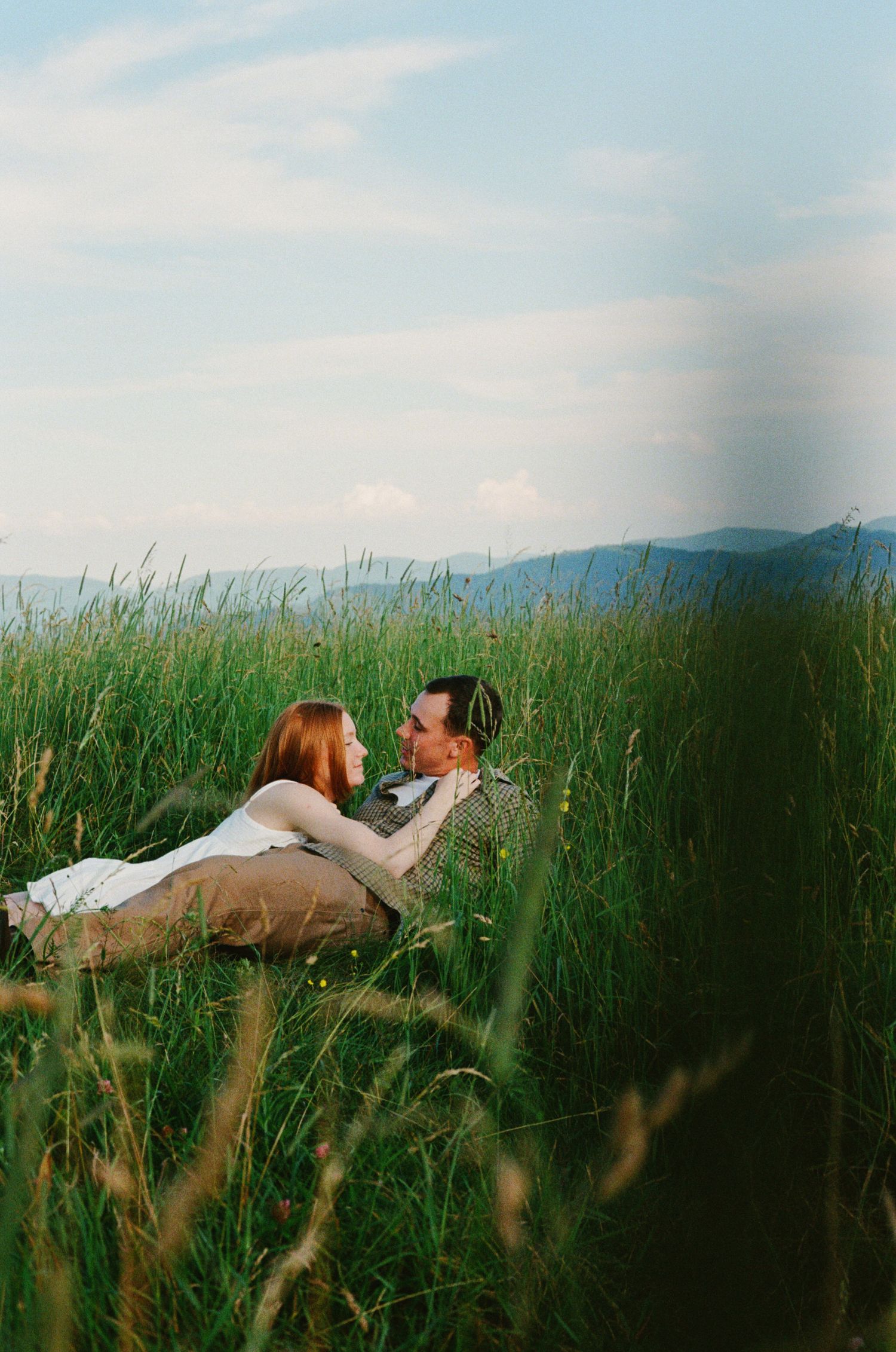 A couple reclines peacefully in a tall grass meadow against a mountain backdrop on a summer afternoon.