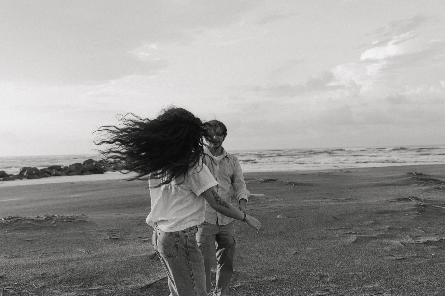 Black and white photo of a couple embracing on a windy beach with waves and dramatic clouds in the background.