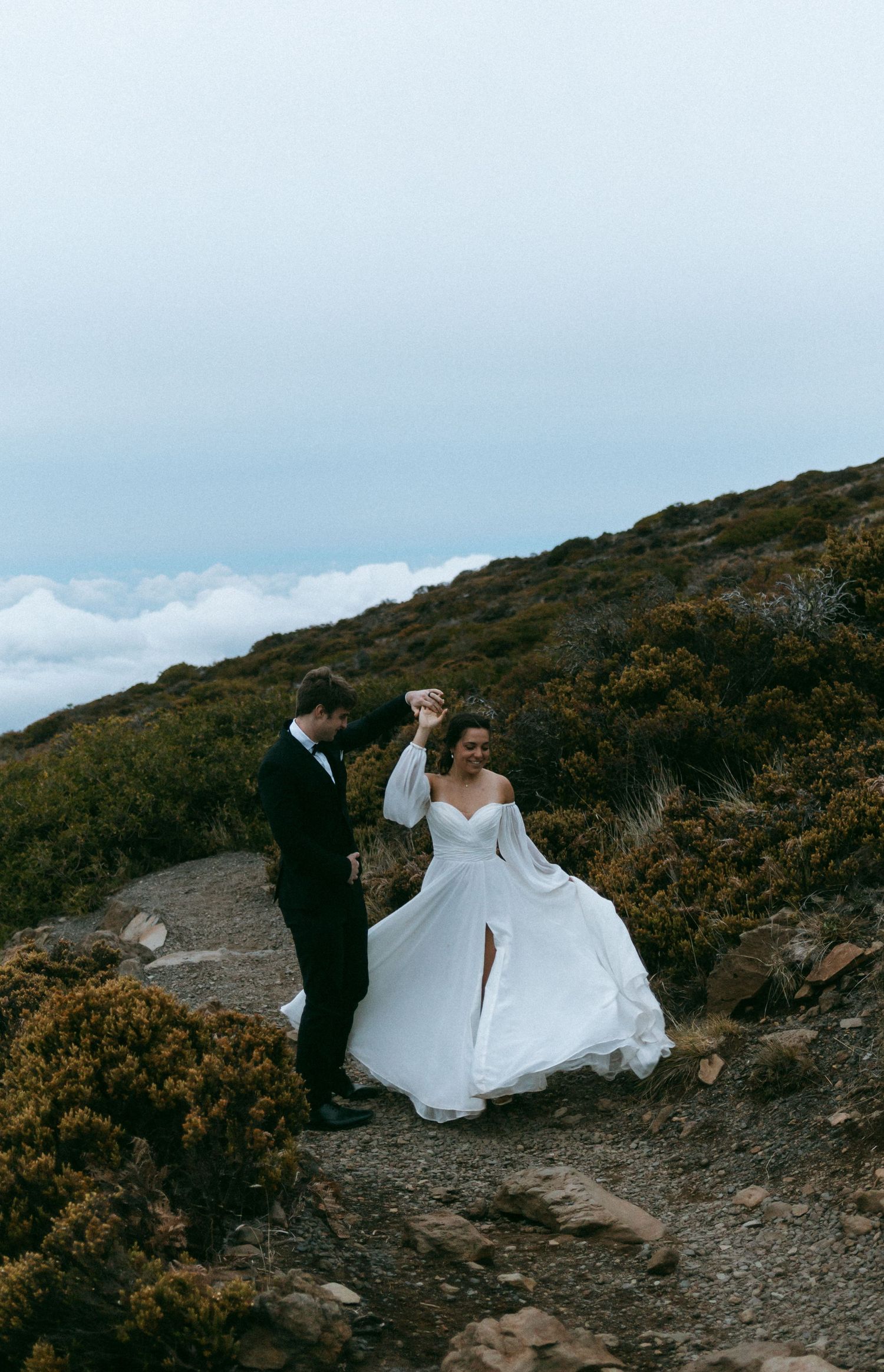 A romantic wedding photoshoot at sunset on a mountain trail with dramatic clouds in the background.