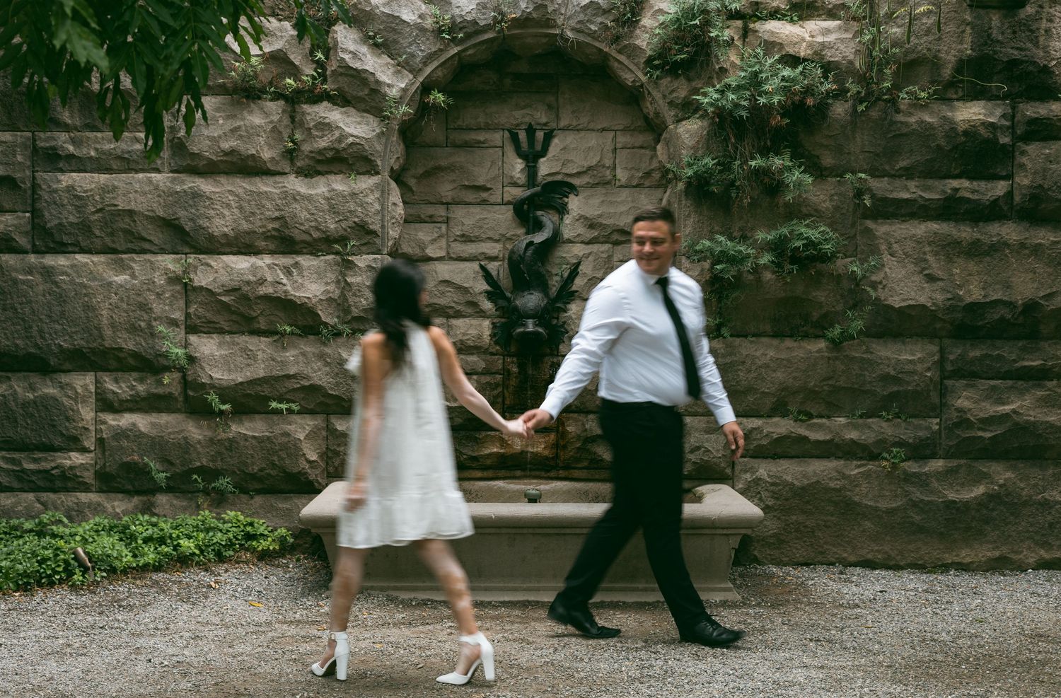 A couple holds hands while walking against a stone wall in a romantic outdoor setting.