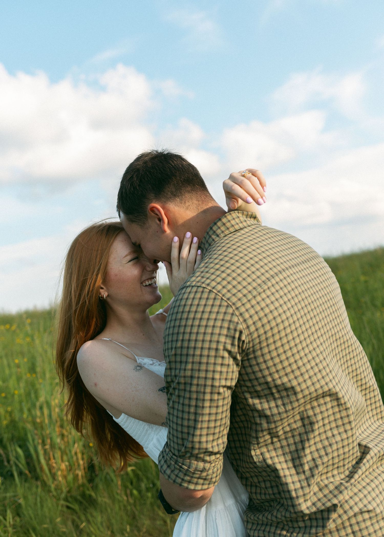 A romantic couple embraces and kisses in a sunlit meadow wearing casual attire against a blue sky background.