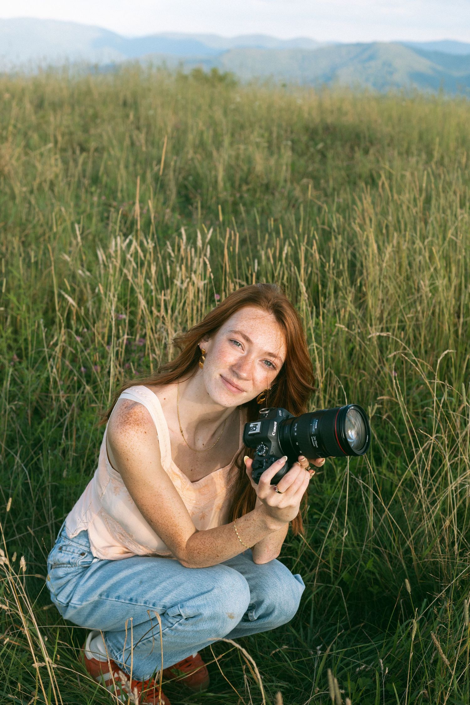 Photographer with professional camera crouches in tall grass with mountains visible in background during golden hour.