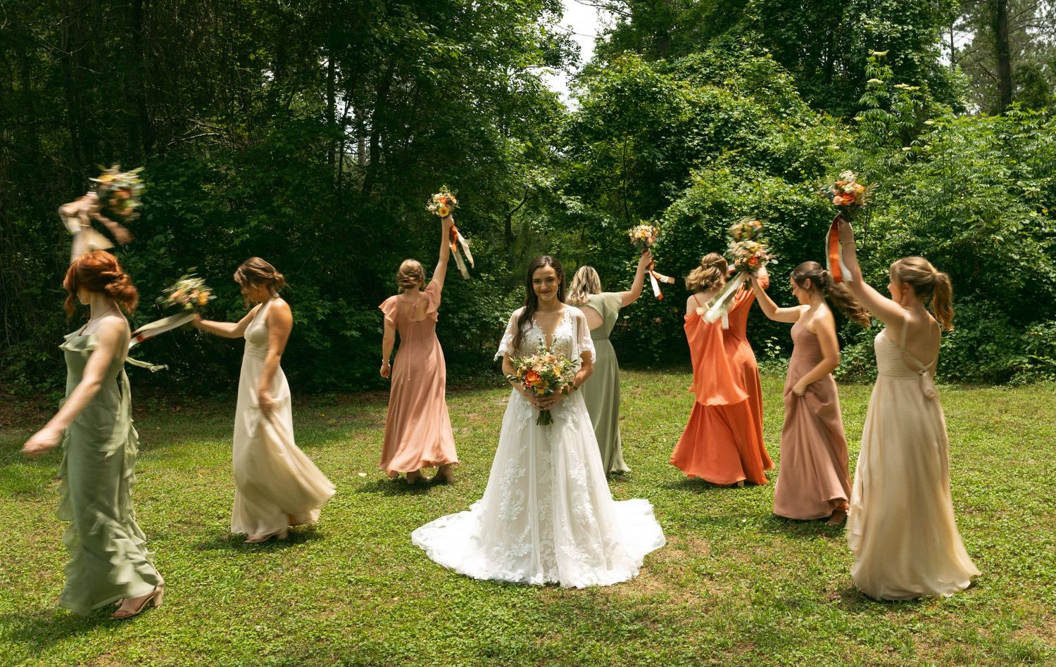 A joyful bridal party in flowing pastel dresses dance in a circle on a lush green lawn surrounded by trees.