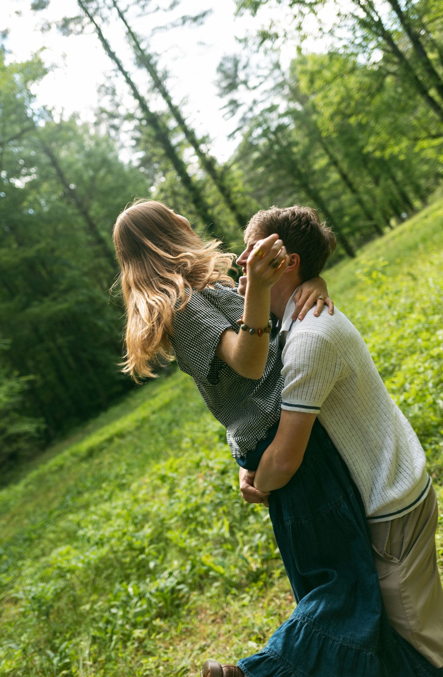 A romantic couple embraces on a grassy hillside surrounded by lush green trees on a sunny summer day.