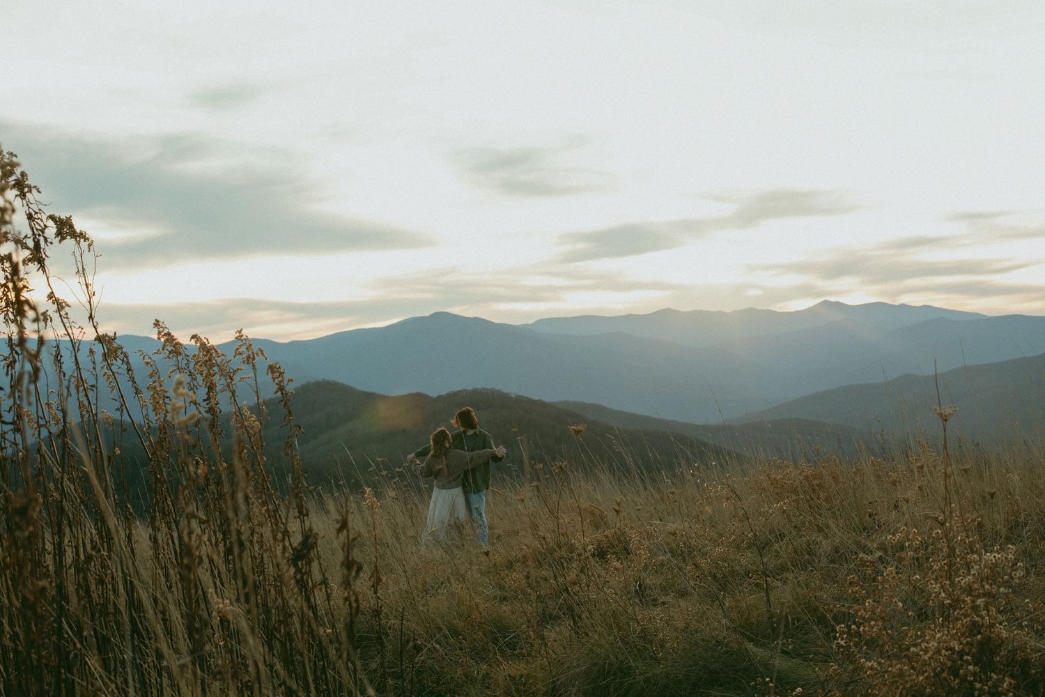 Hiker stands on a mountain ridge at sunset overlooking hazy mountain ranges and tall grass in the foreground.