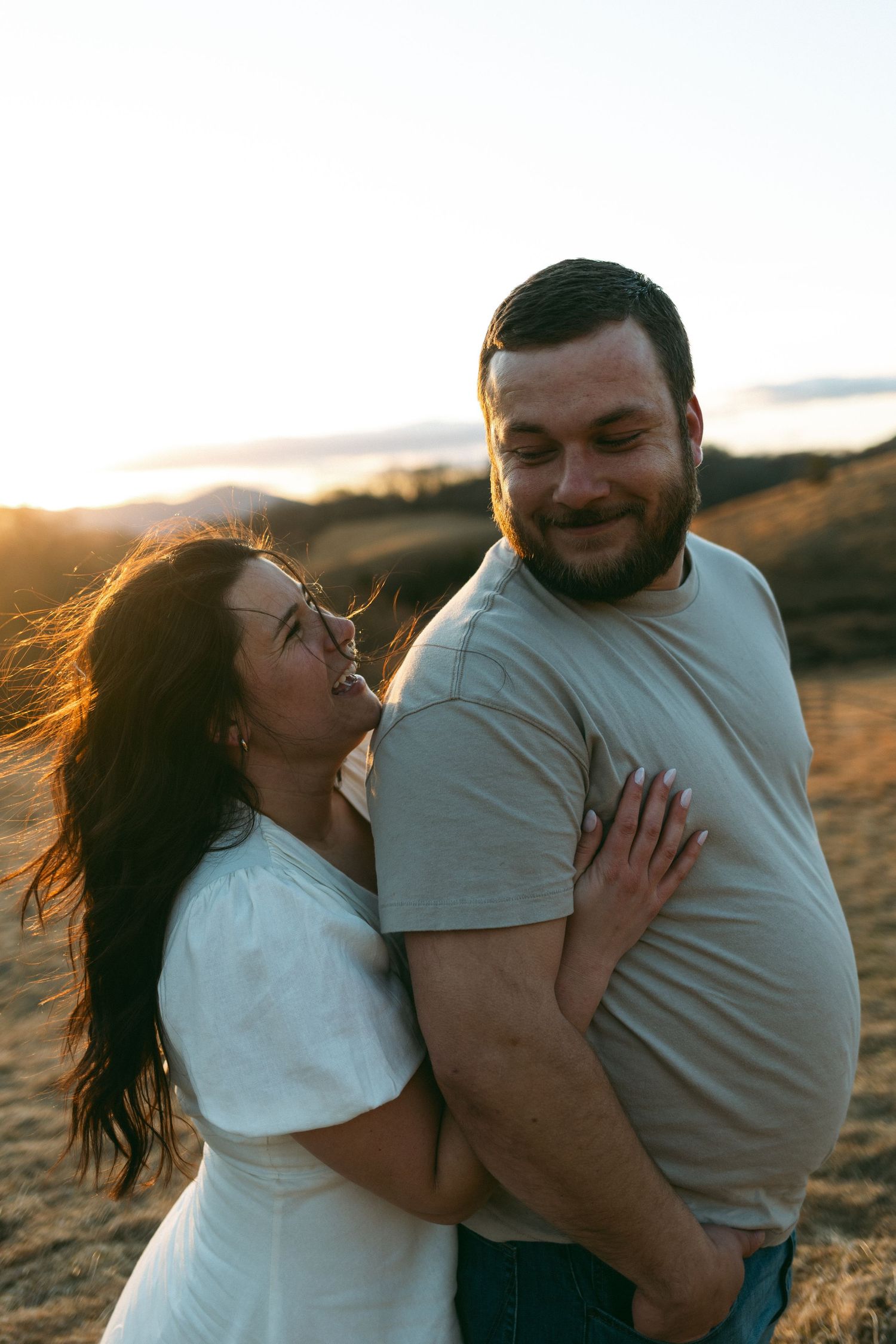 A couple embraces and laughs together during golden hour sunset on a rocky hillside.