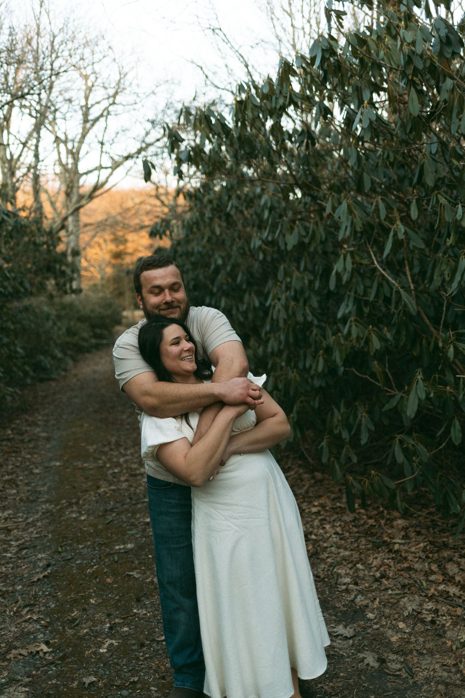 A couple embraces on a nature trail surrounded by green foliage and trees at dusk.