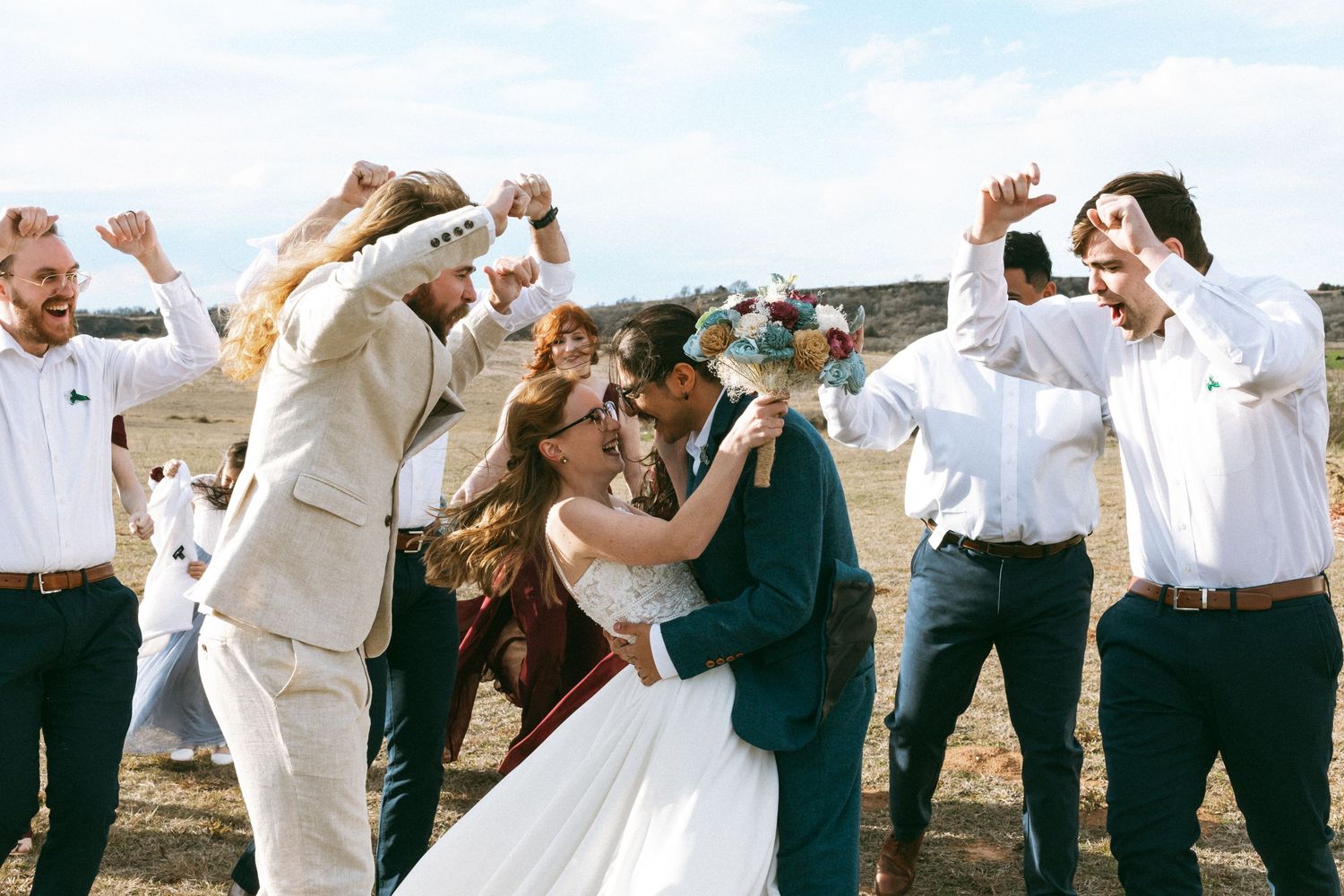 Wedding party celebrates with raised arms during outdoor ceremony in rustic setting with natural lighting and joyful energy.