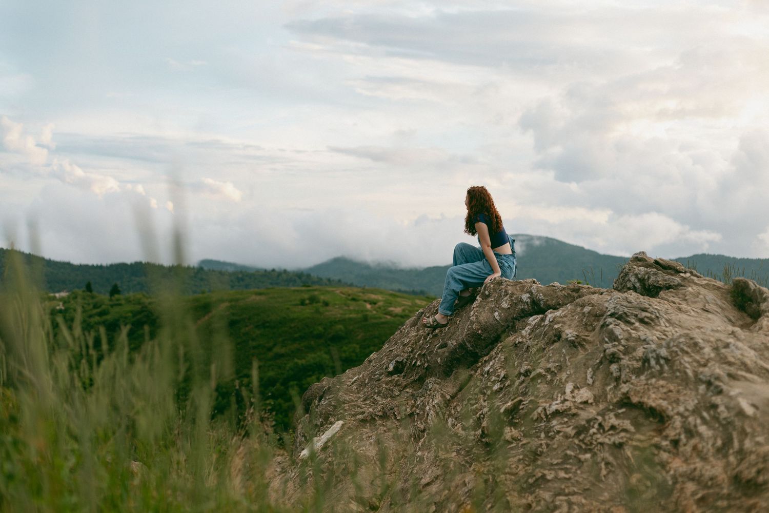 Someone sits peacefully on a hilltop overlooking a scenic mountain landscape on a cloudy summer day.