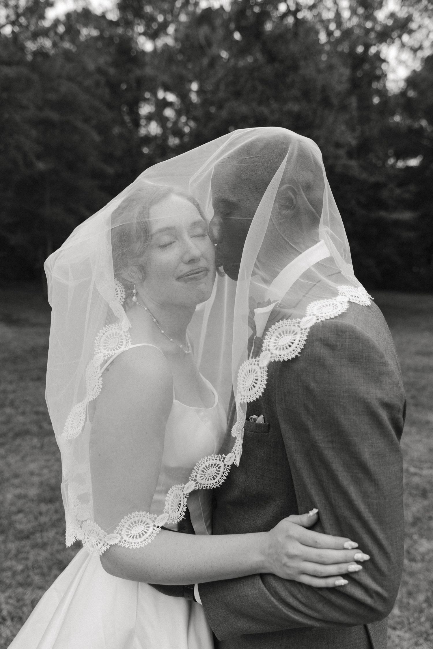 A romantic black and white wedding photo showing a couple embracing under an elegant lace-trimmed veil in an outdoor setting.
