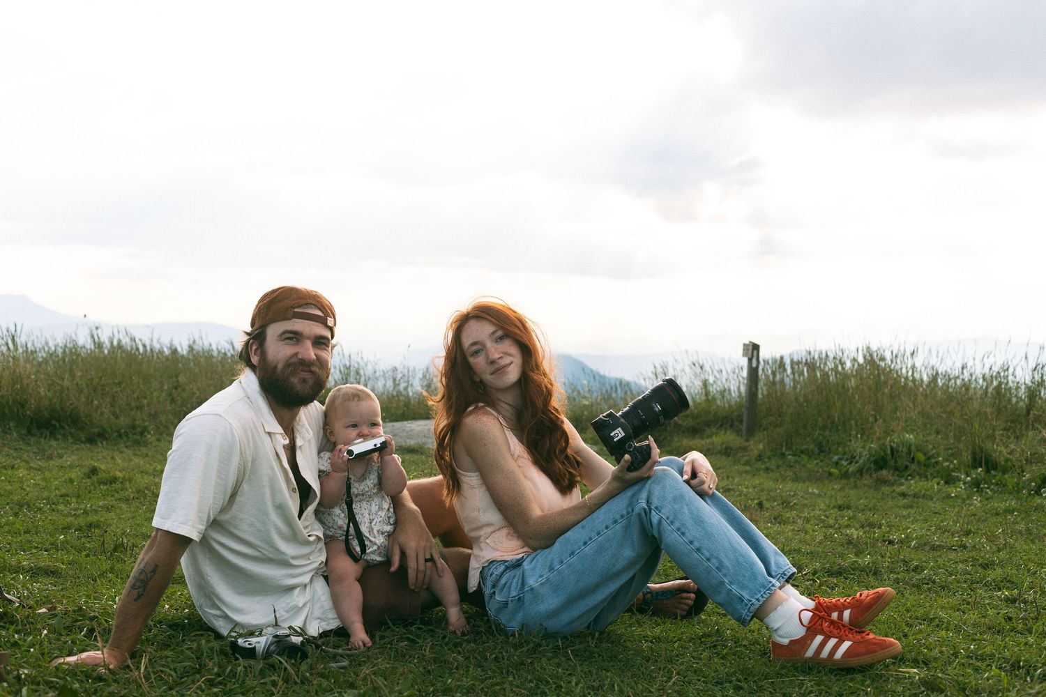 A family relaxes together on grassy hillside during golden hour in casual summer attire.