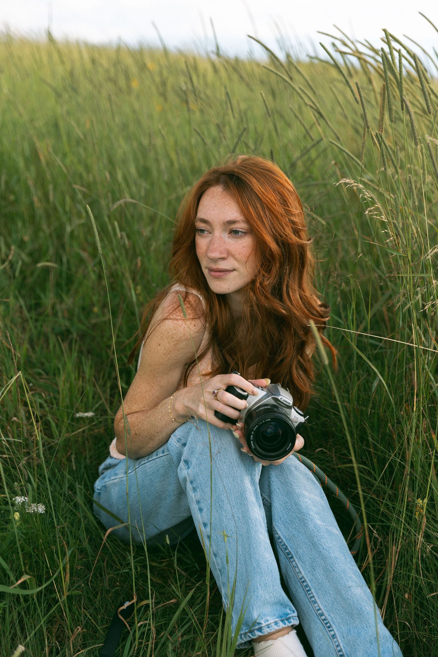 Photographer with long red hair sits in tall grass field with a vintage camera at golden hour.