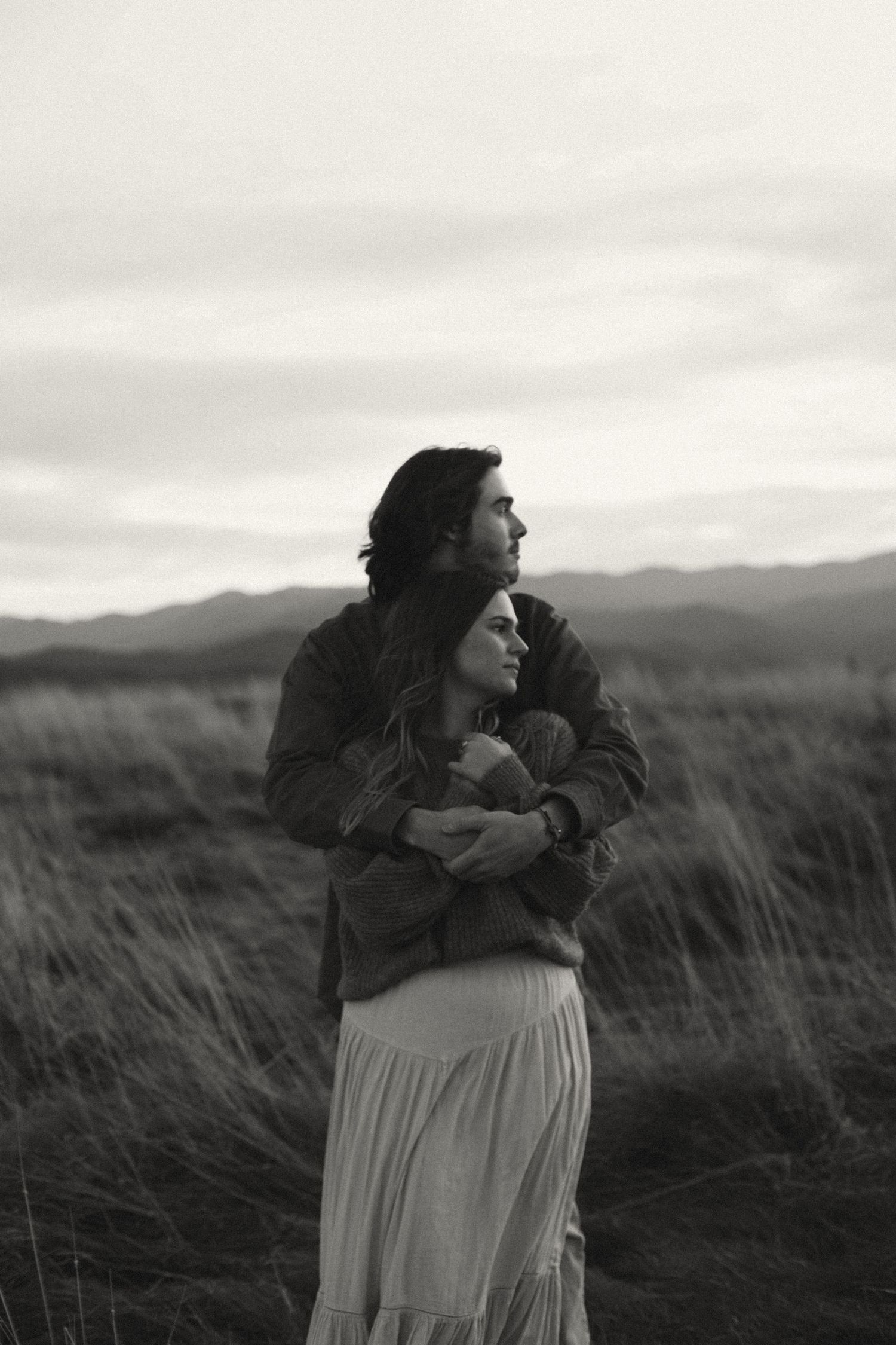 A romantic black and white photograph of a couple embracing in a windswept field with mountains in the background.