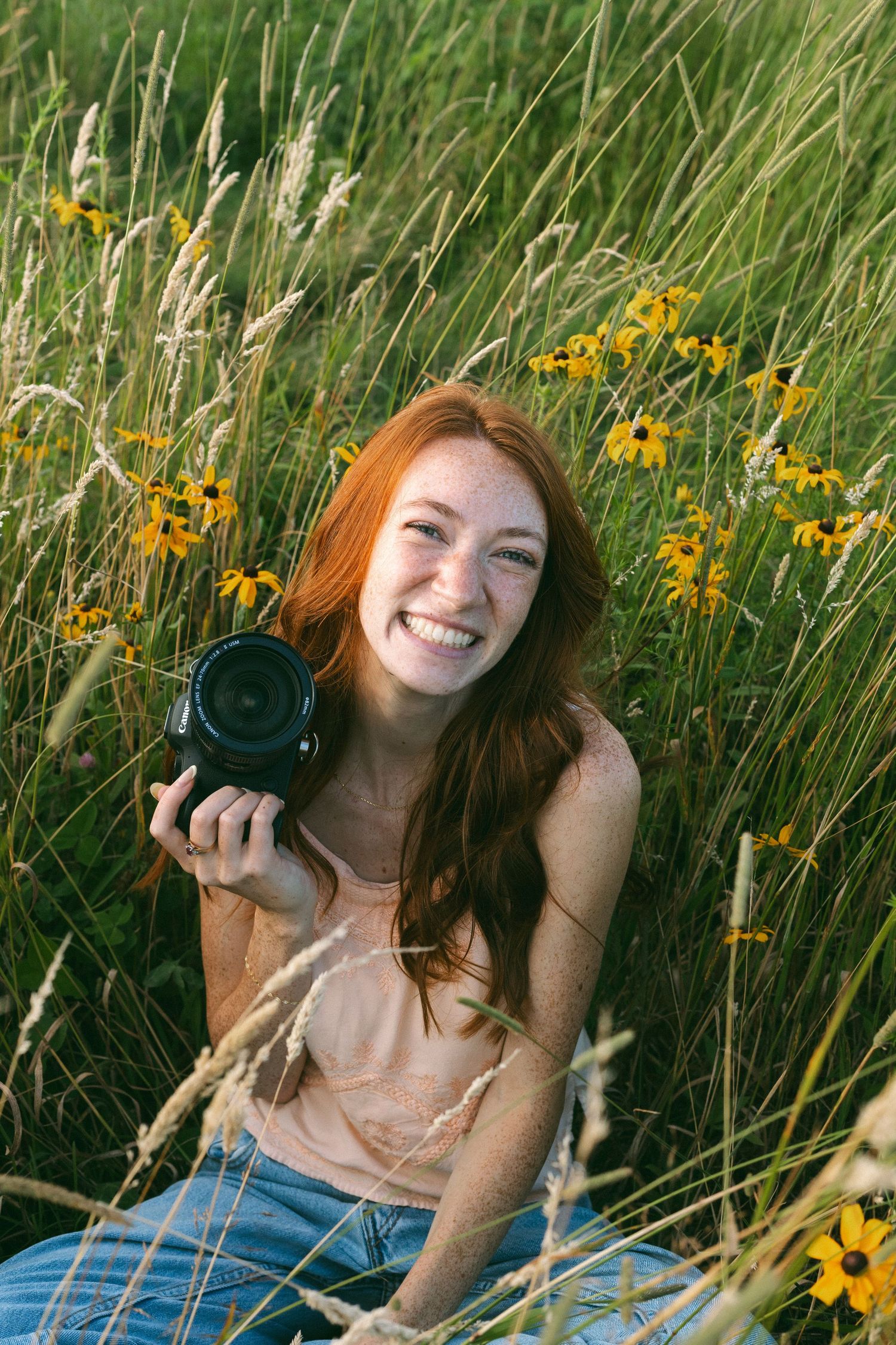 A photographer lies in a field of tall grass and yellow wildflowers on a sunny summer day with a vintage camera.