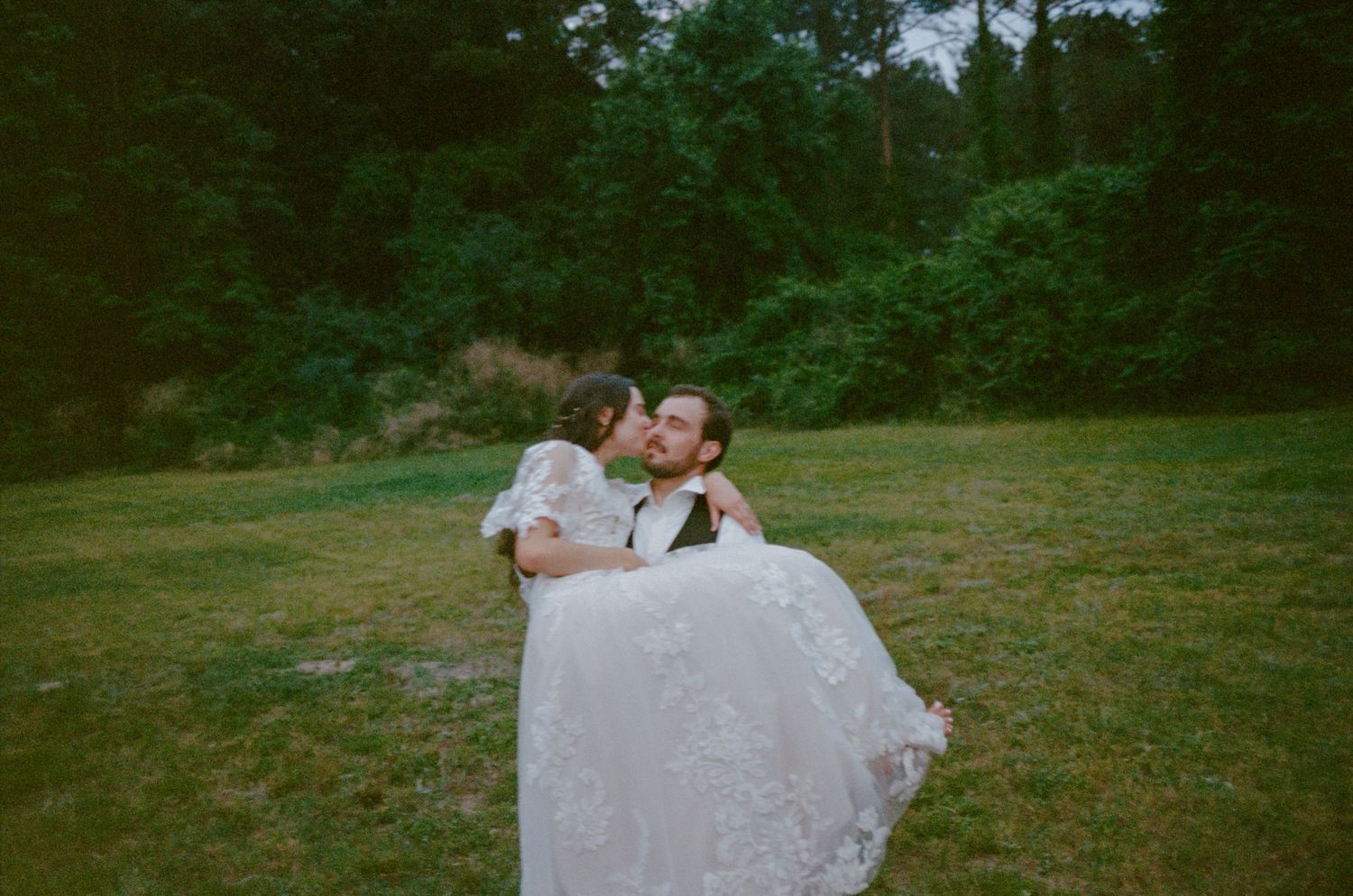 A couple shares an intimate wedding moment on a grassy field with forest trees in the background.