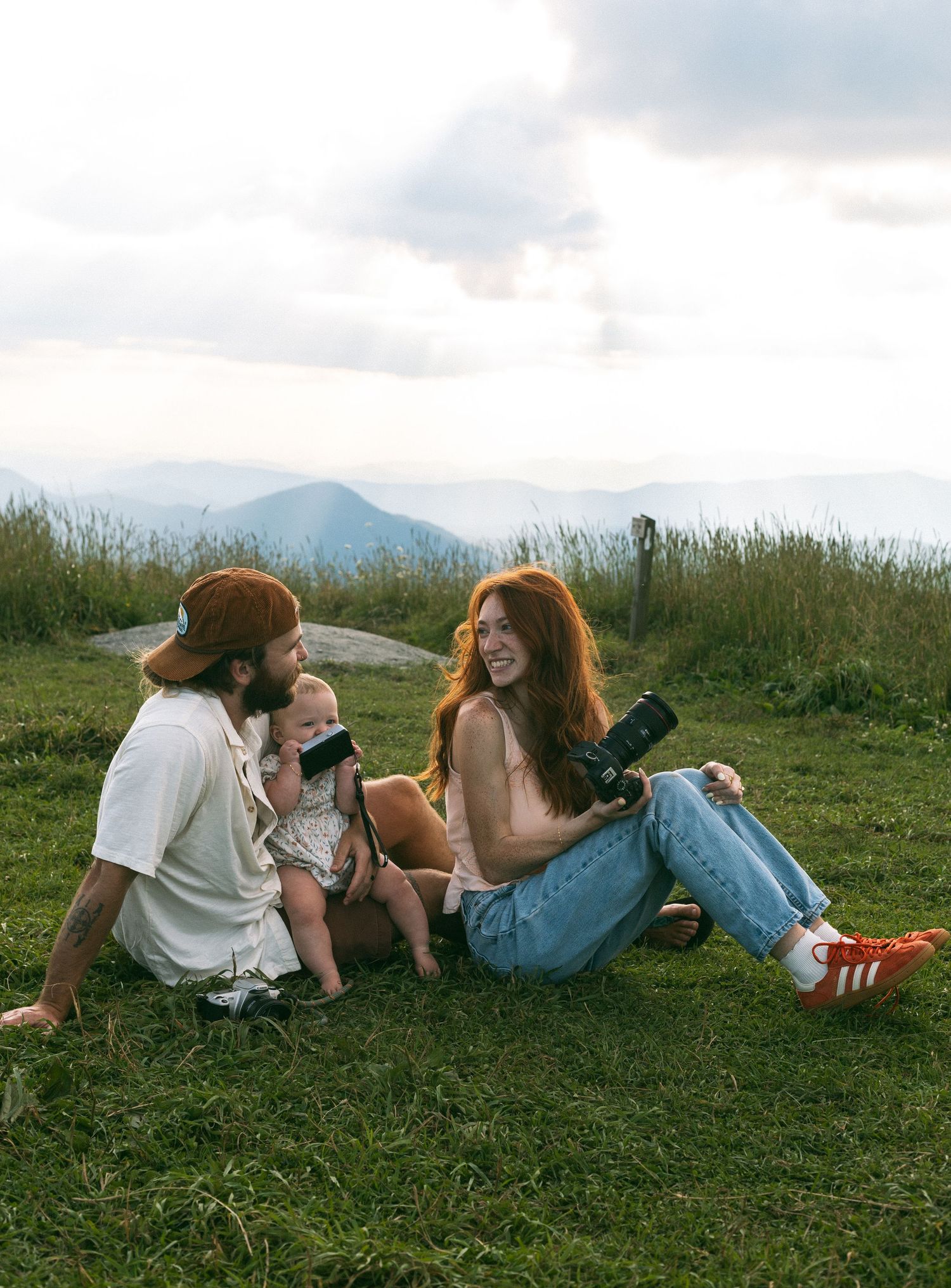 Three friends relaxing on grassy hilltop with mountains in background on a cloudy day.