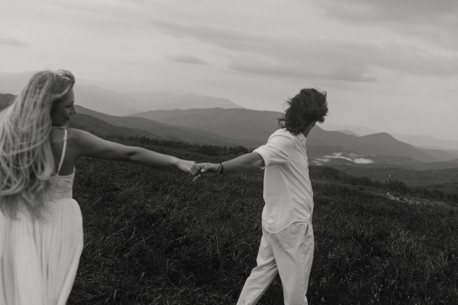 Two silhouettes holding hands on a hilltop overlook with mountains in the distance against a cloudy sky.