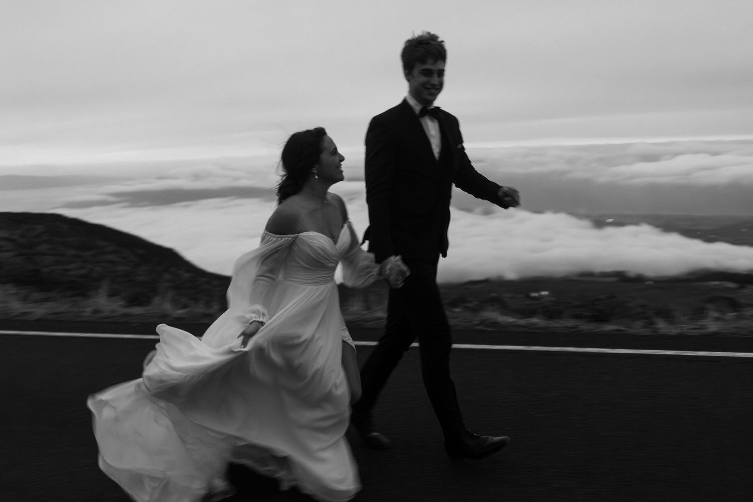 Couple in wedding attire walking along a mountain road with dramatic clouds below at sunset in black and white.