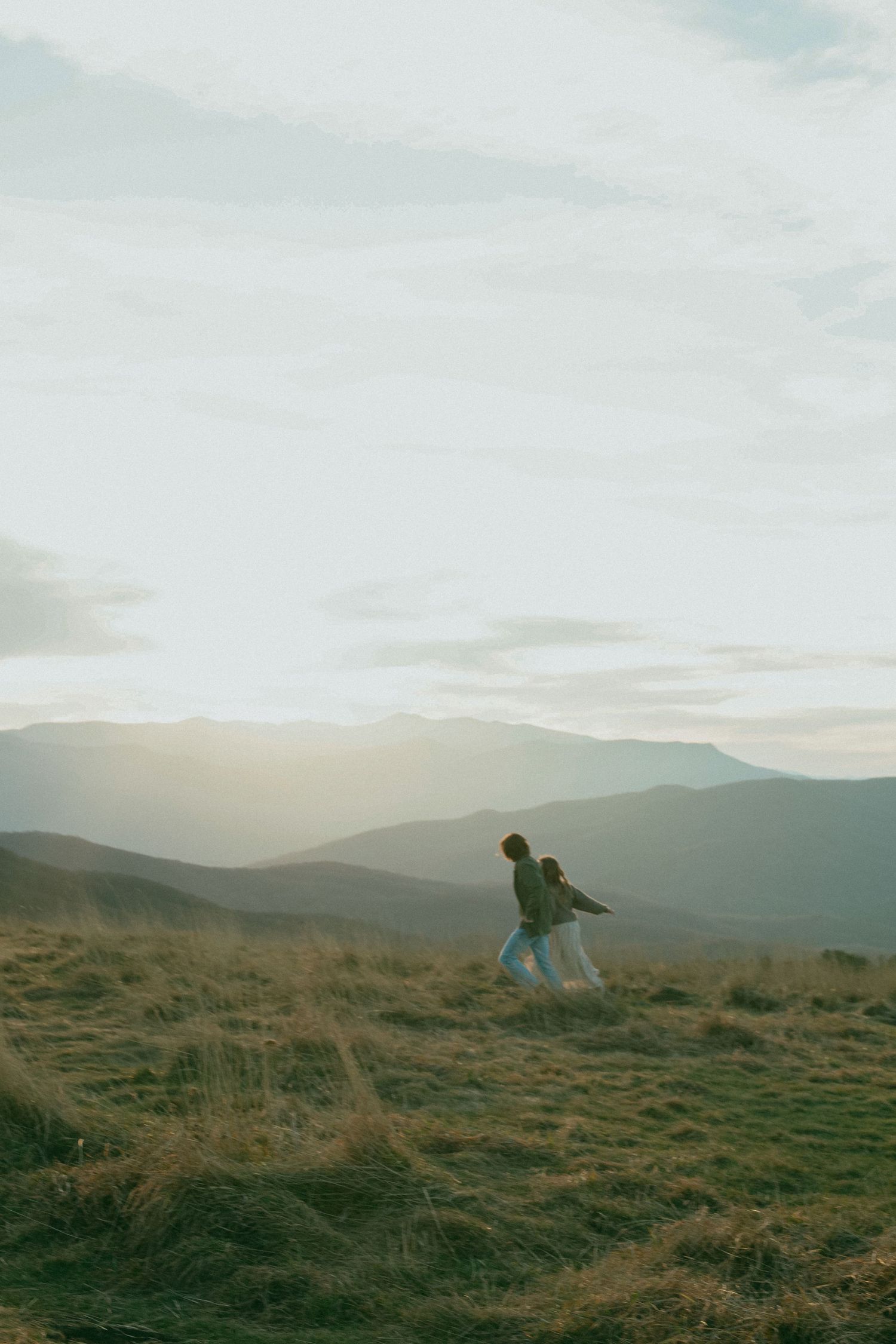 Silhouettes of two people walking together on a hilltop against misty mountain ranges during golden hour.