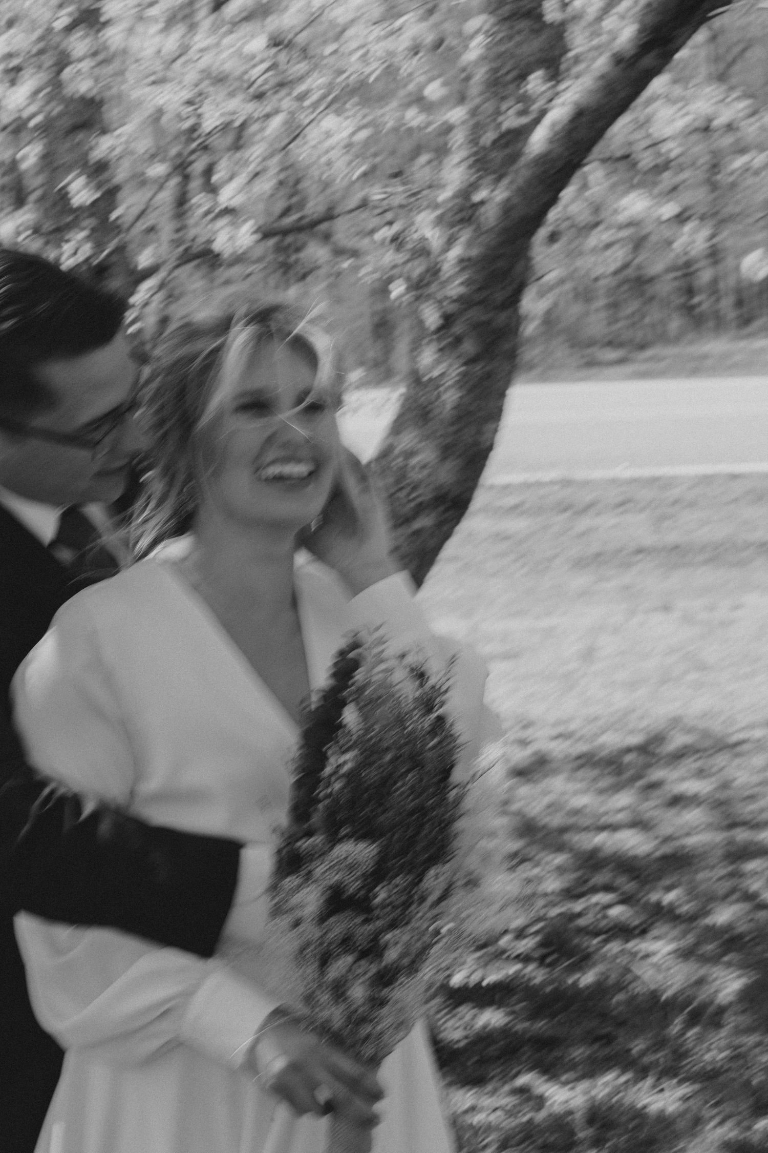 A joyful black and white wedding photo shows a couple sharing a candid moment together outdoors by a tree.