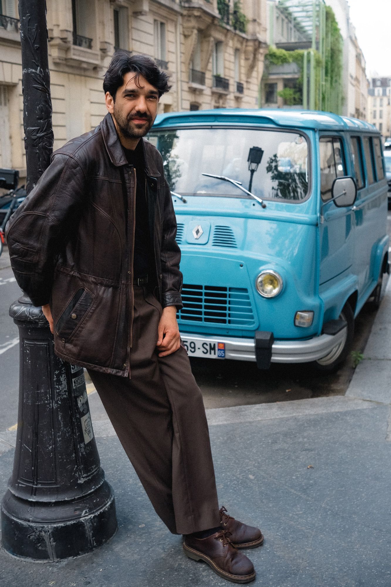 Person in dark clothing poses near classic blue cars parked on a Parisian street.
