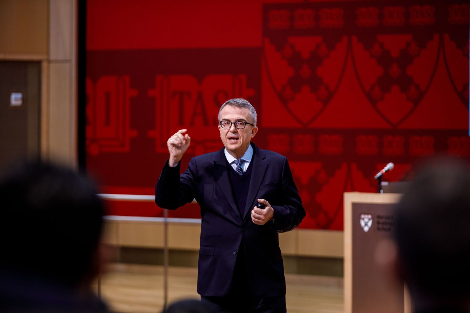 A speaker gestures while presenting at a Harvard University conference in a red-themed auditorium.