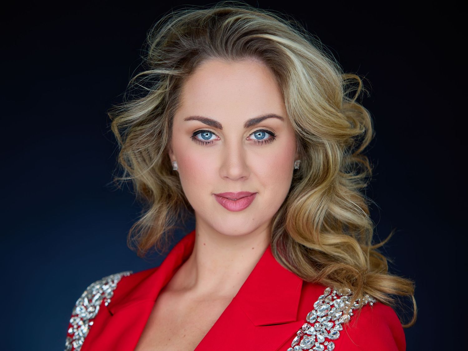 Professional headshot of a blonde woman with blue eyes wearing a red jeweled blazer against a dark background.