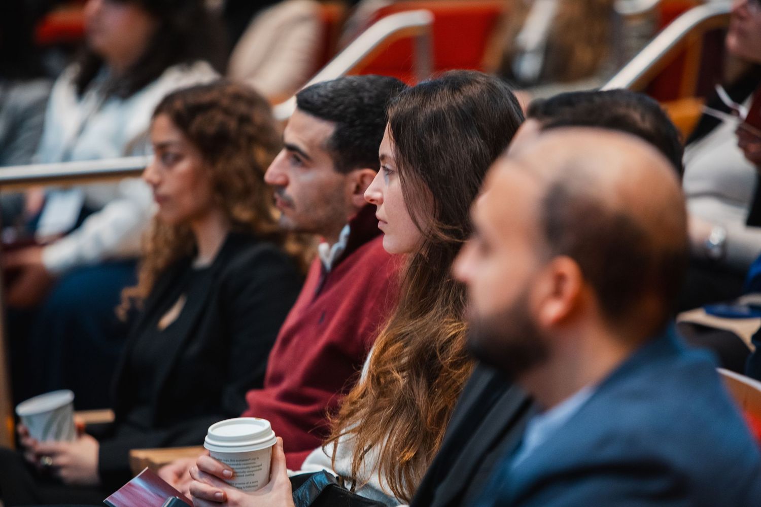 Engaged audience members listen attentively at an academic conference, one holding a coffee cup.