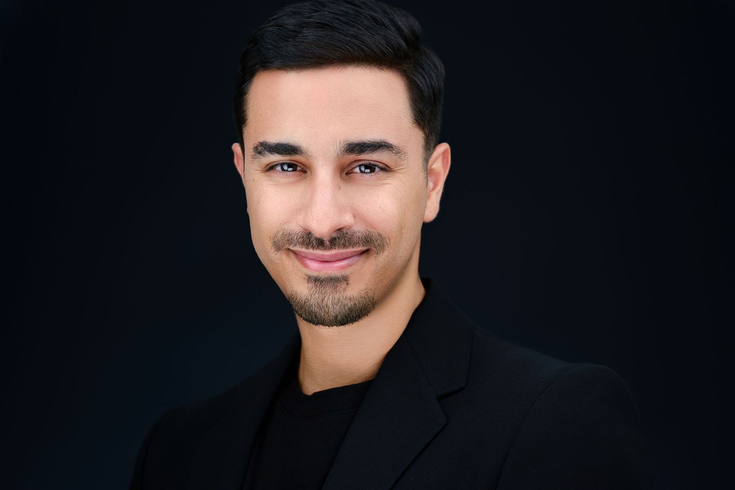 Professional headshot of a young man in a black blazer smiling confidently against a dark background.