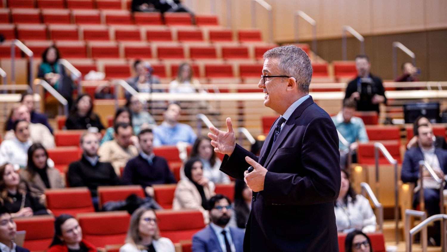 A dynamic speaker addresses an attentive audience in a modern red-seated university auditorium.