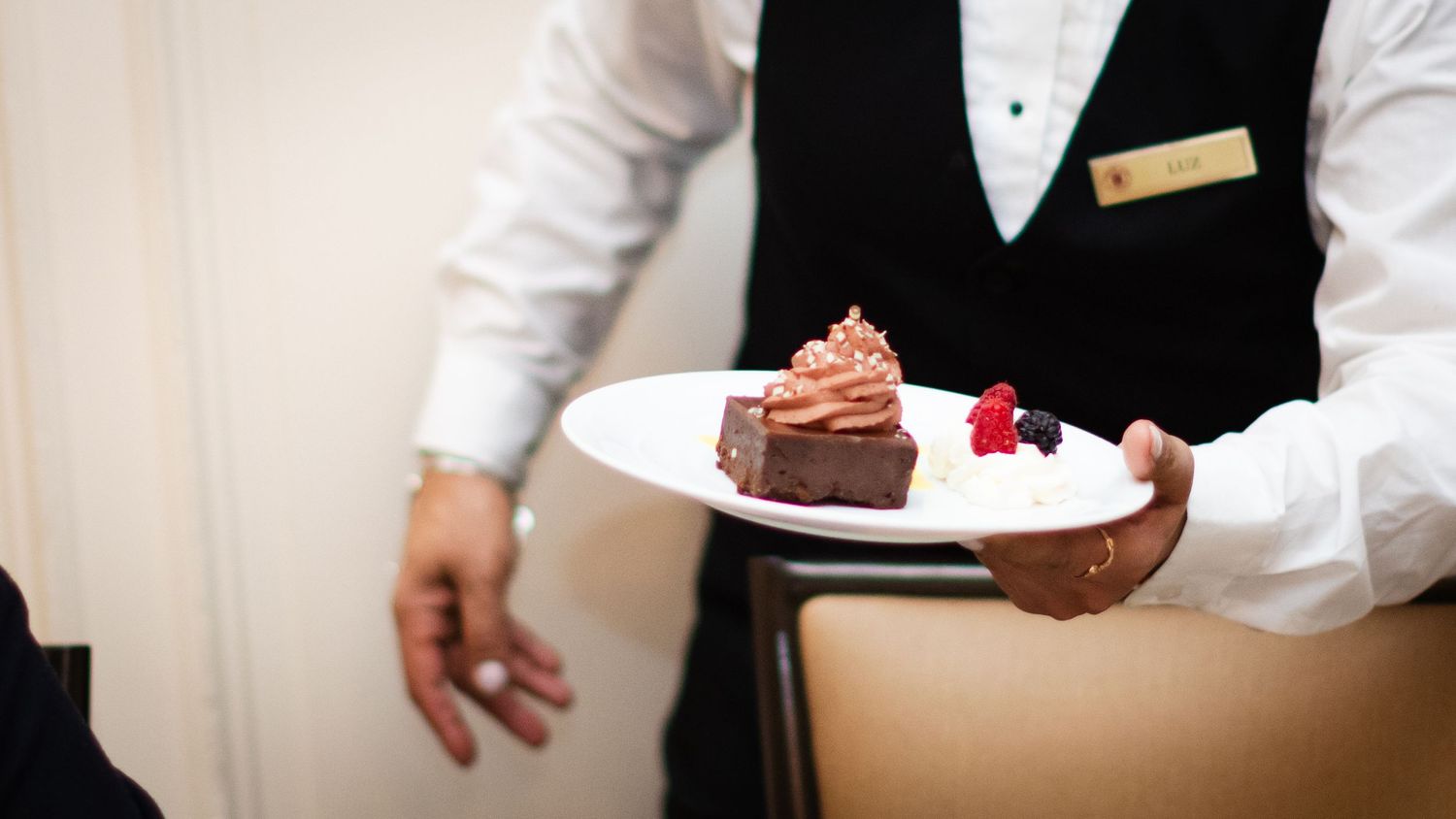 Formally dressed waiter presenting a chocolate dessert with whipped cream and berries.