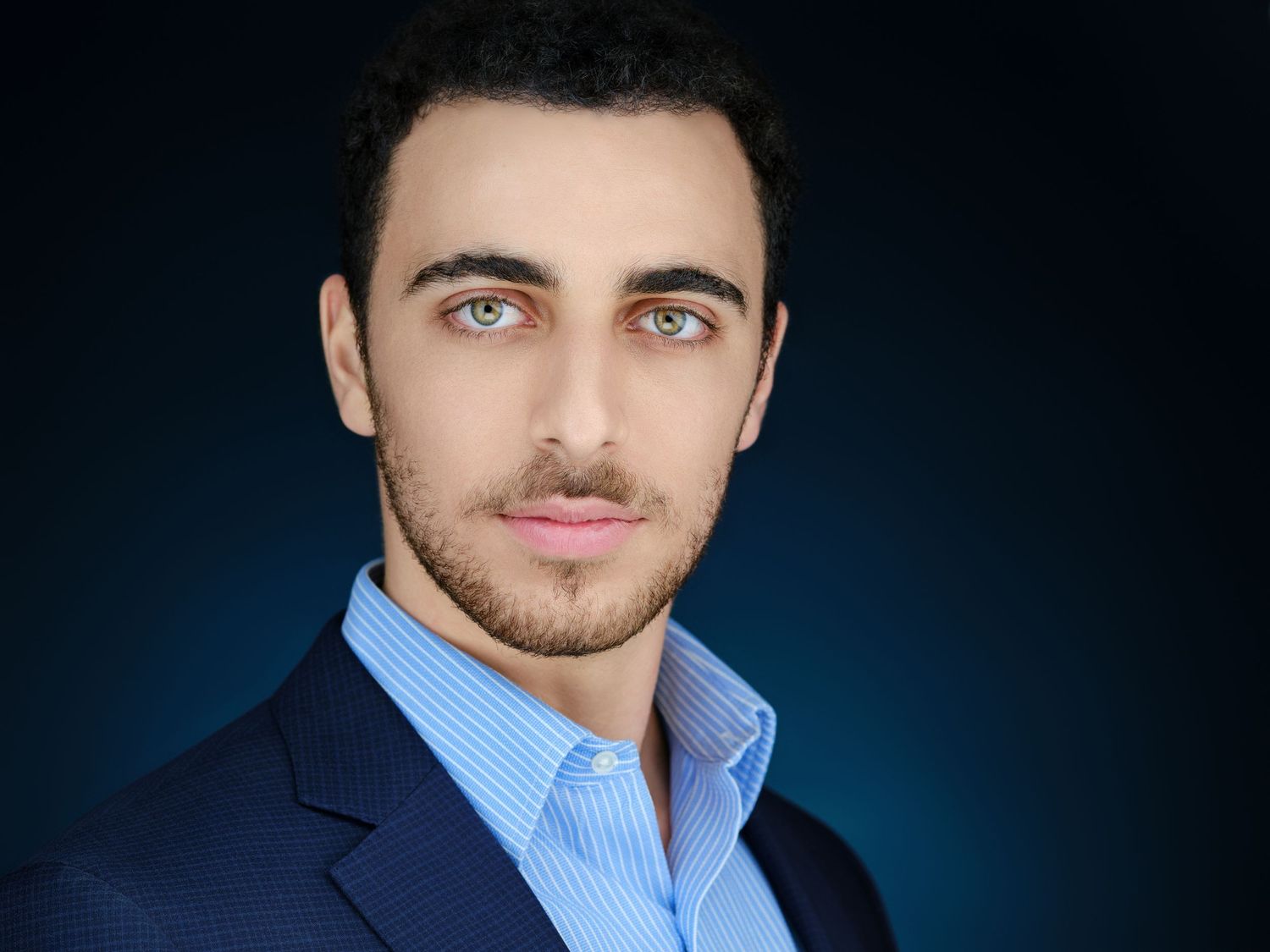 Professional headshot of a young man in a navy suit and light blue shirt against a dark blue background.