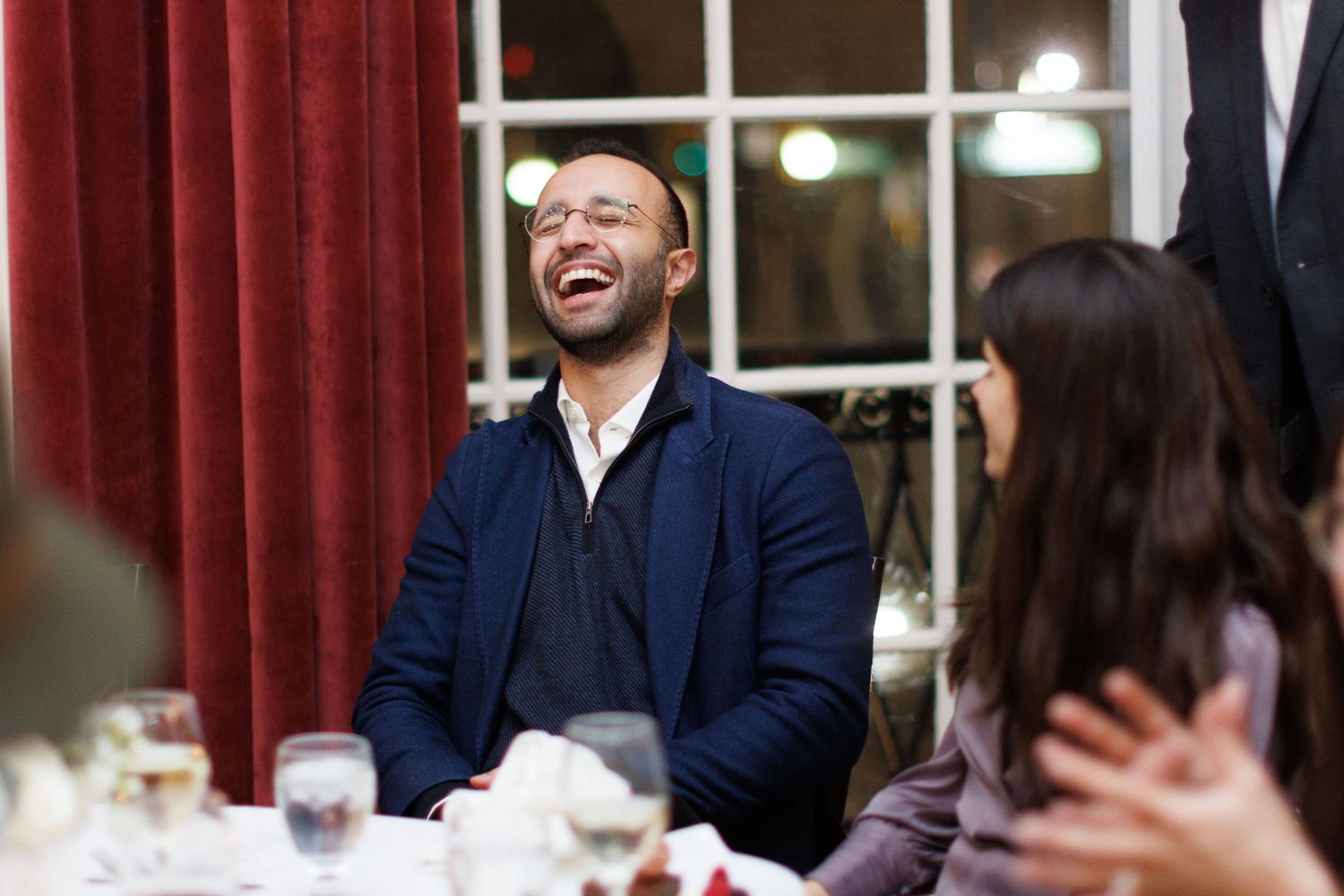 Man in navy blazer laughing at a sophisticated dinner event with red curtain backdrop.
