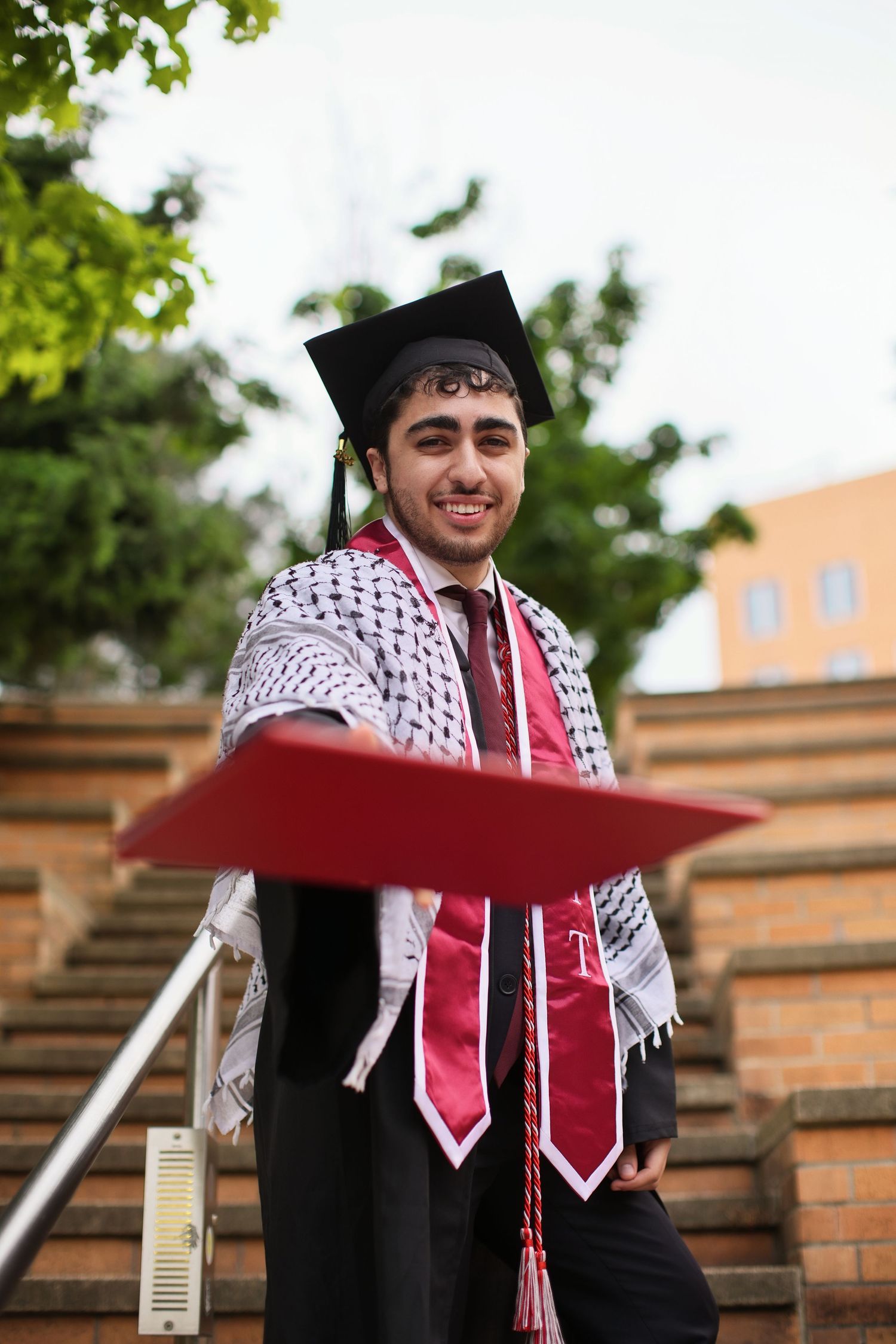 Smiling graduate in cap and gown with keffiyeh holds diploma on outdoor steps.