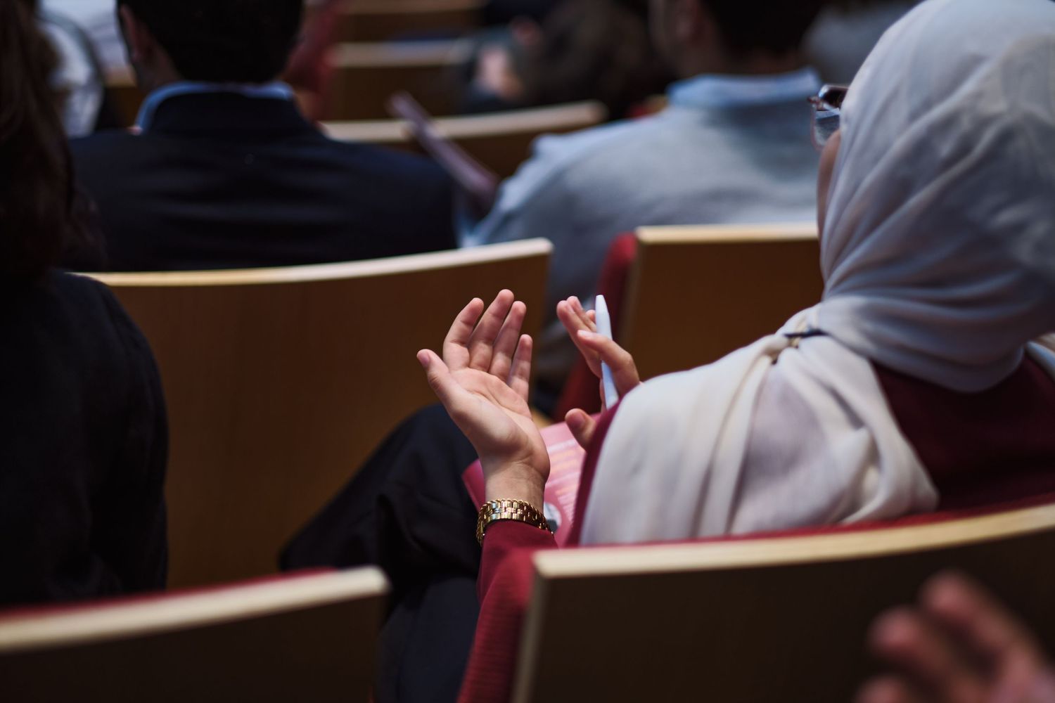 An audience member in a hijab applauds during a conference presentation in a tiered auditorium.