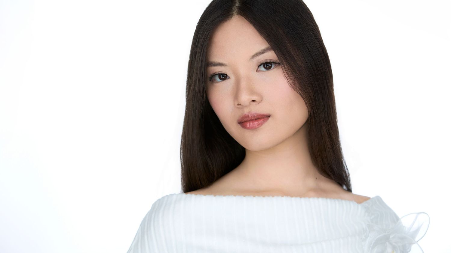 Young Asian woman with straight dark hair wearing a white off-shoulder top, posing against a white background.