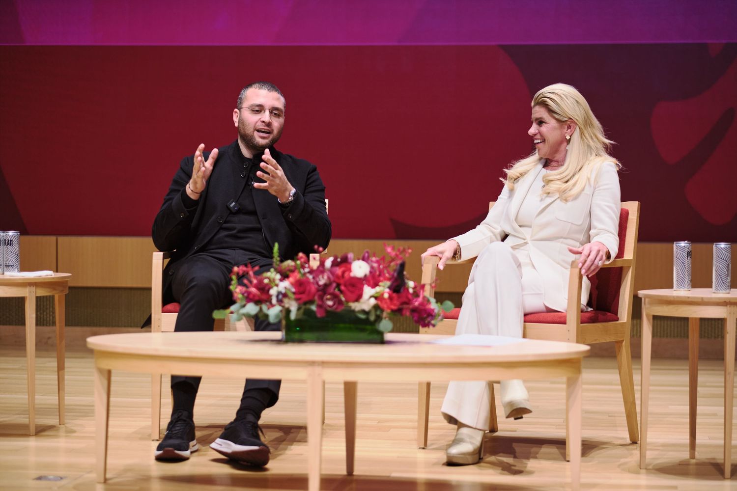 A man and woman engage in a panel discussion seated on wooden chairs with a floral centerpiece.