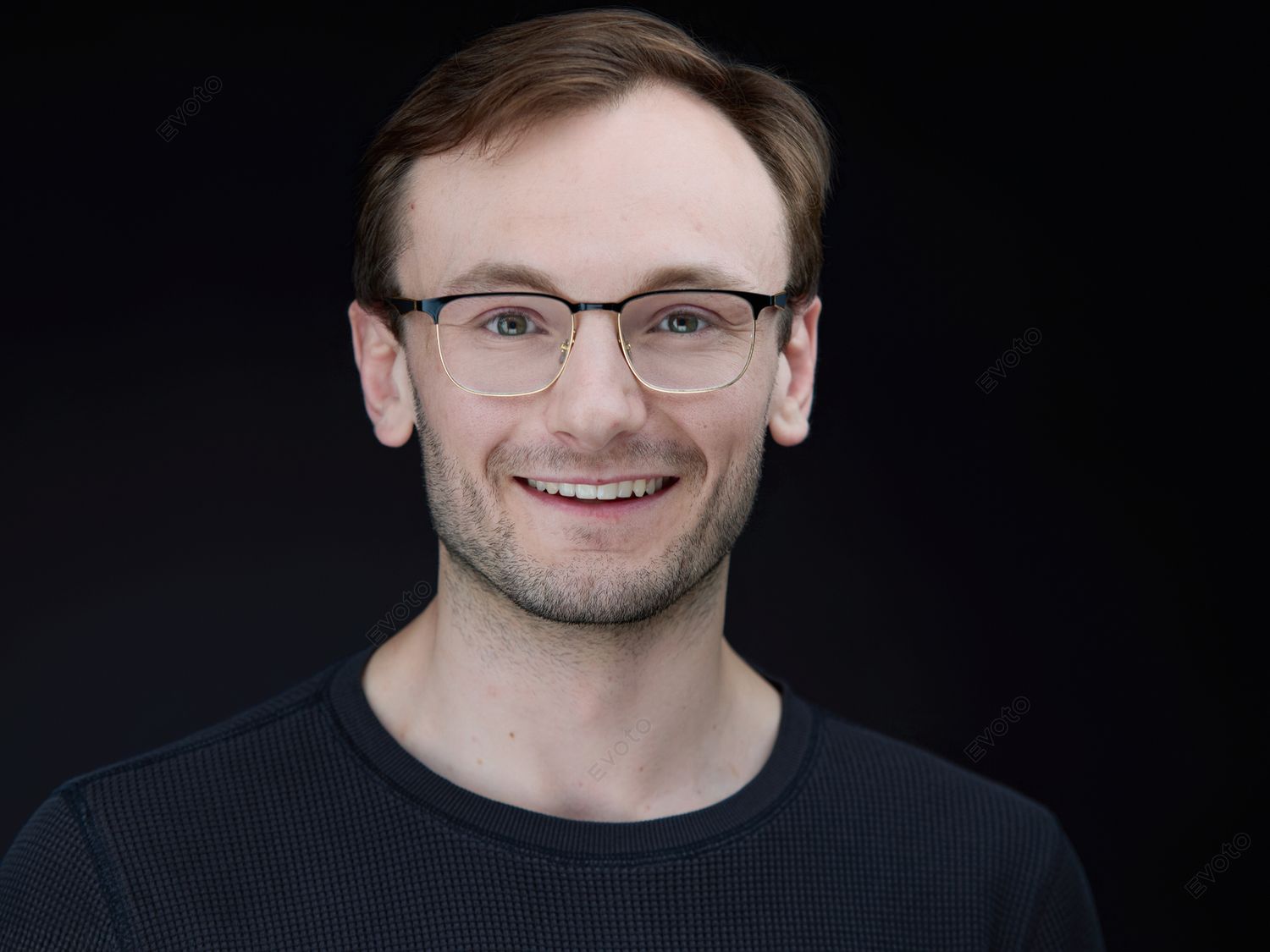 Smiling young man with glasses wearing a dark crew-neck shirt against a black background in a professional headshot.