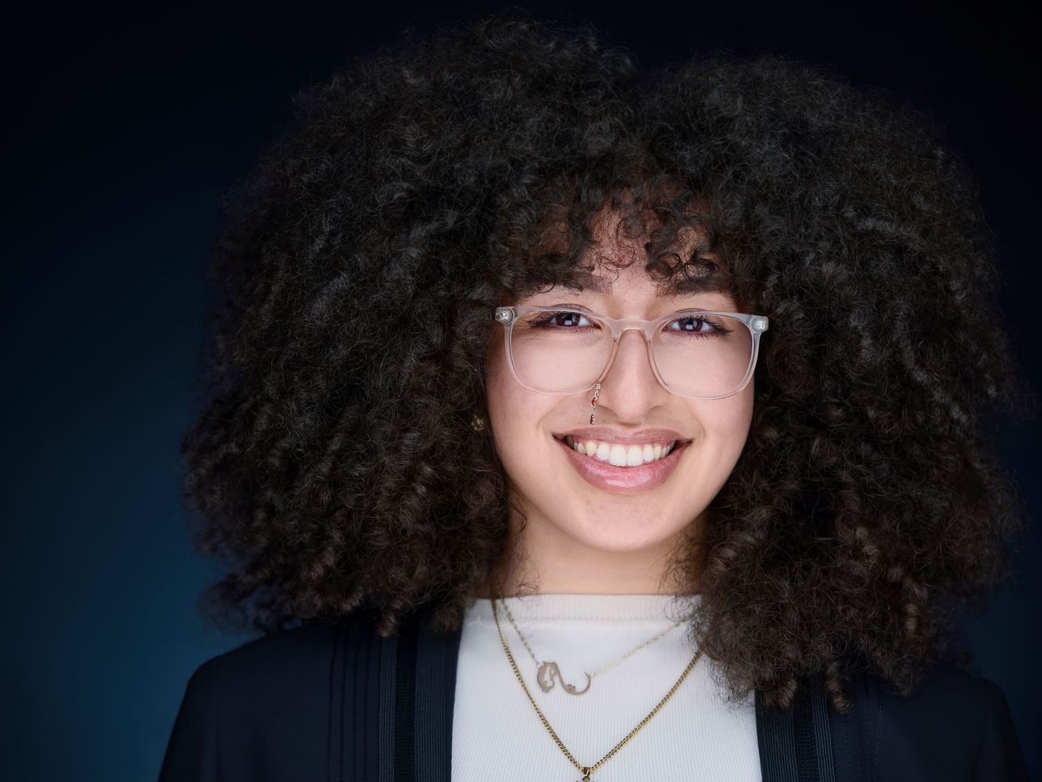 Smiling young woman with curly hair and clear glasses wearing a navy blazer against a dark blue background.