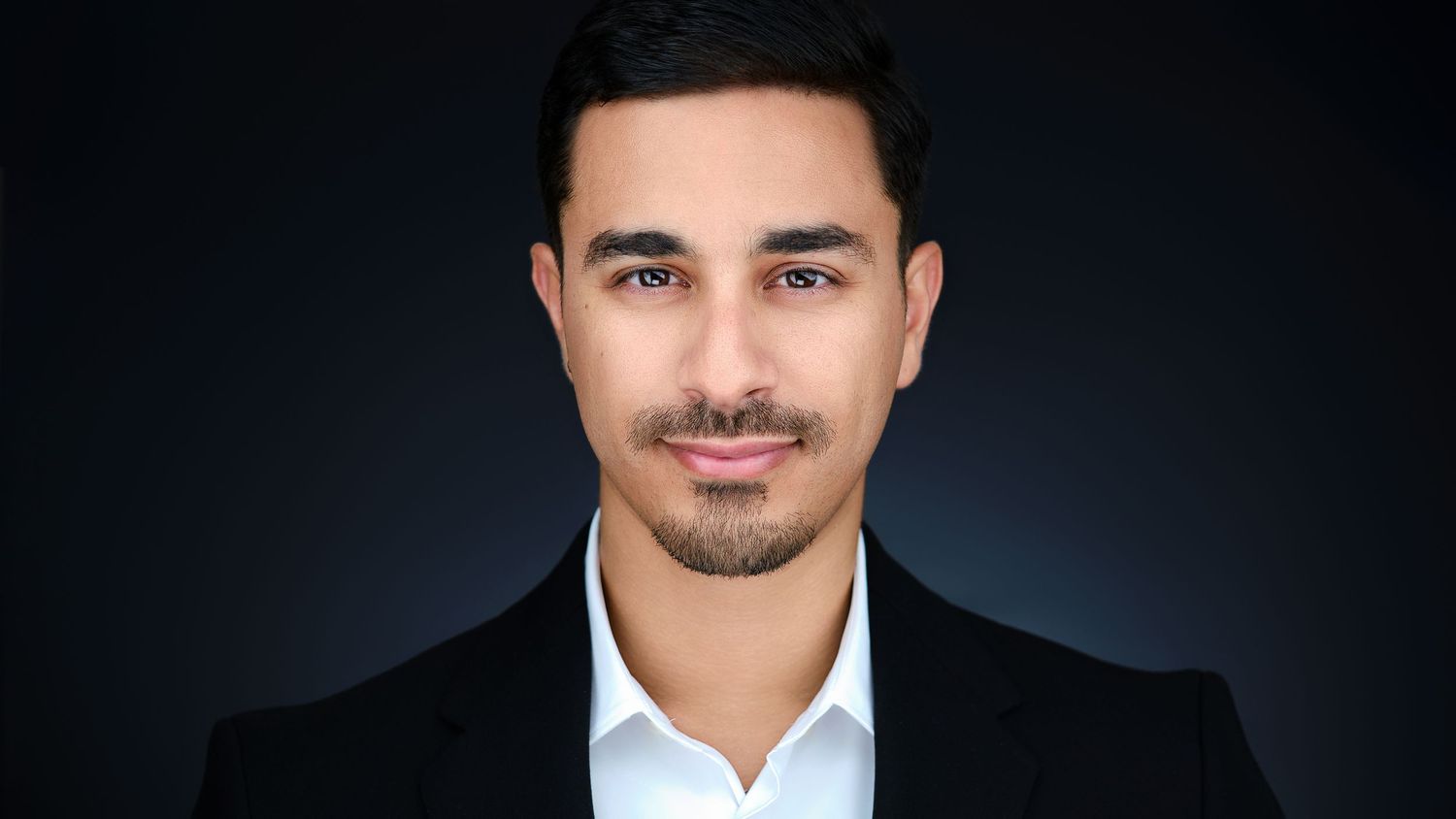 Professional headshot of young man in black blazer and white shirt on dark background.