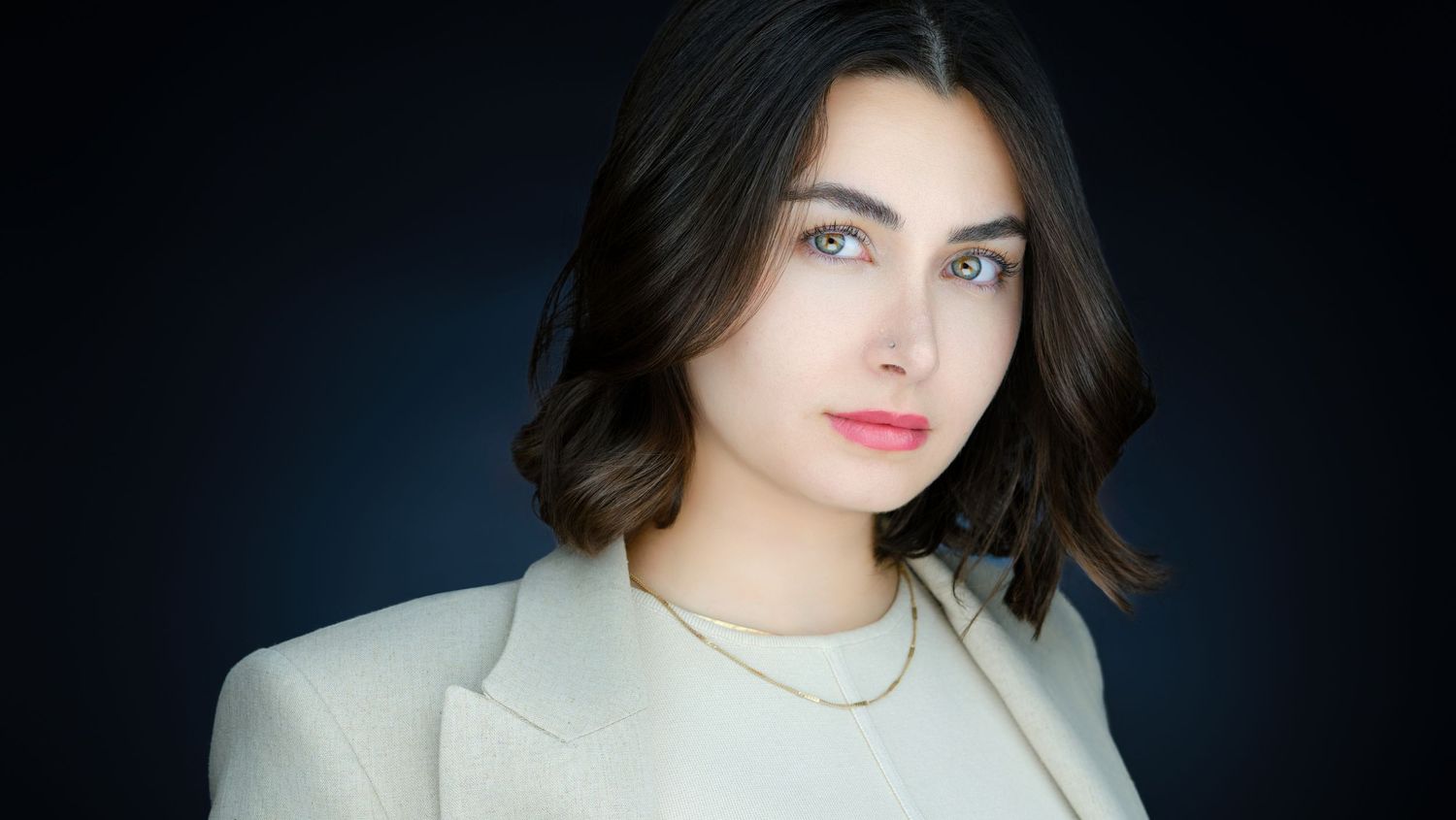 Young woman with dark bob hair wearing a white blazer and gold necklace poses against a dark background.