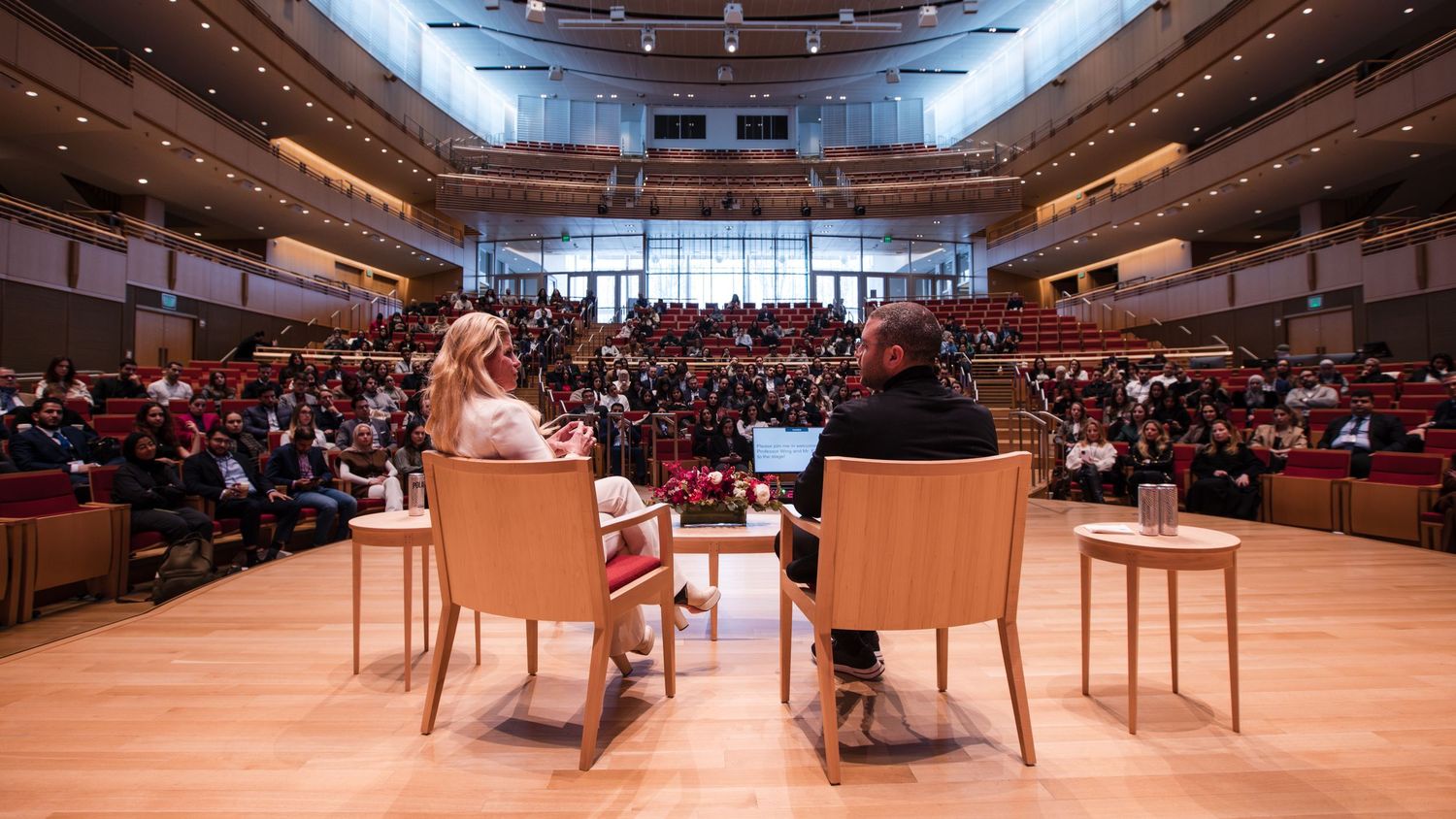 Two speakers seated on stage during a keynote presentation in a packed concert hall.