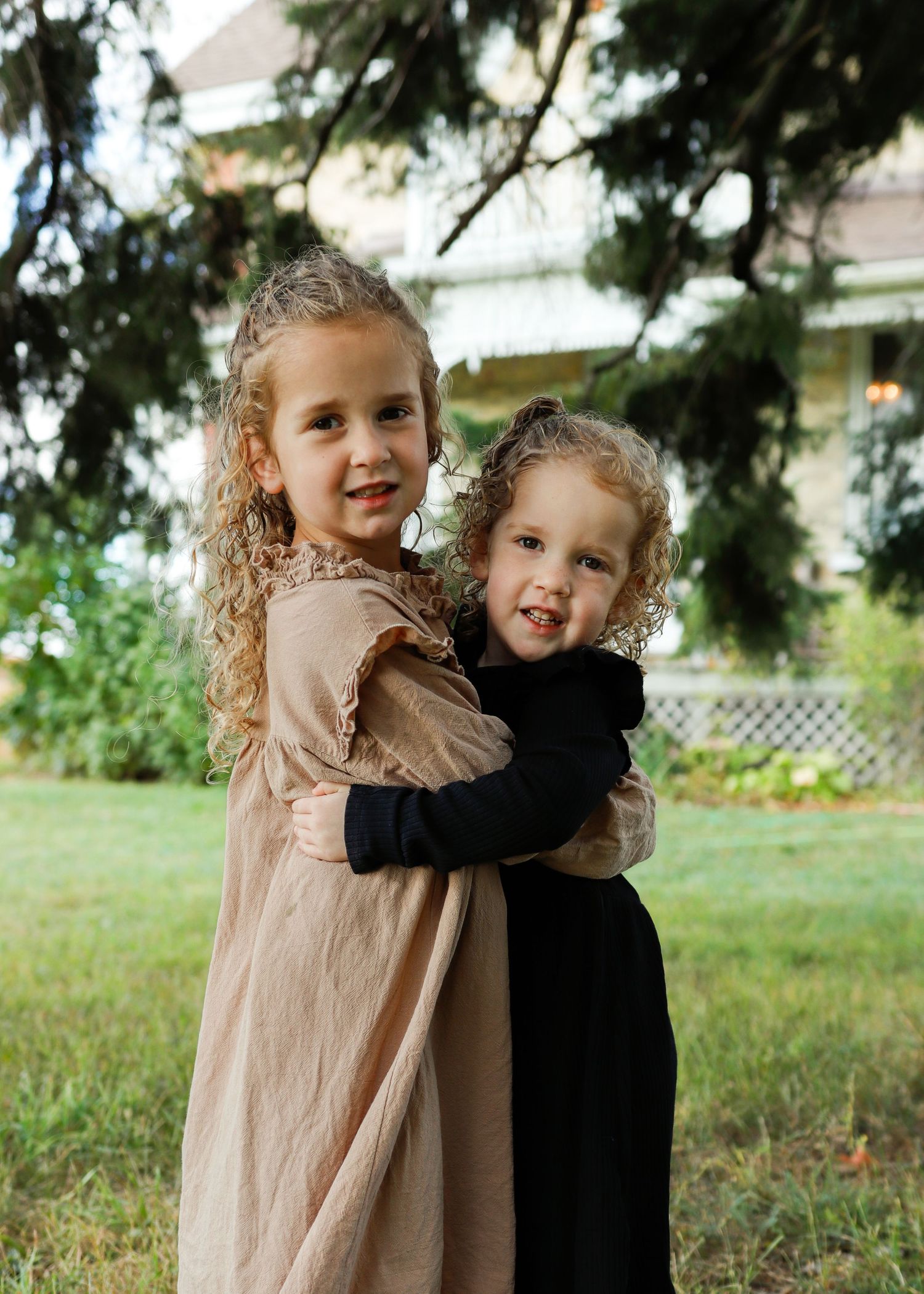 Two young children in beige and black outfits share a sweet embrace while playing outdoors in a grassy area.