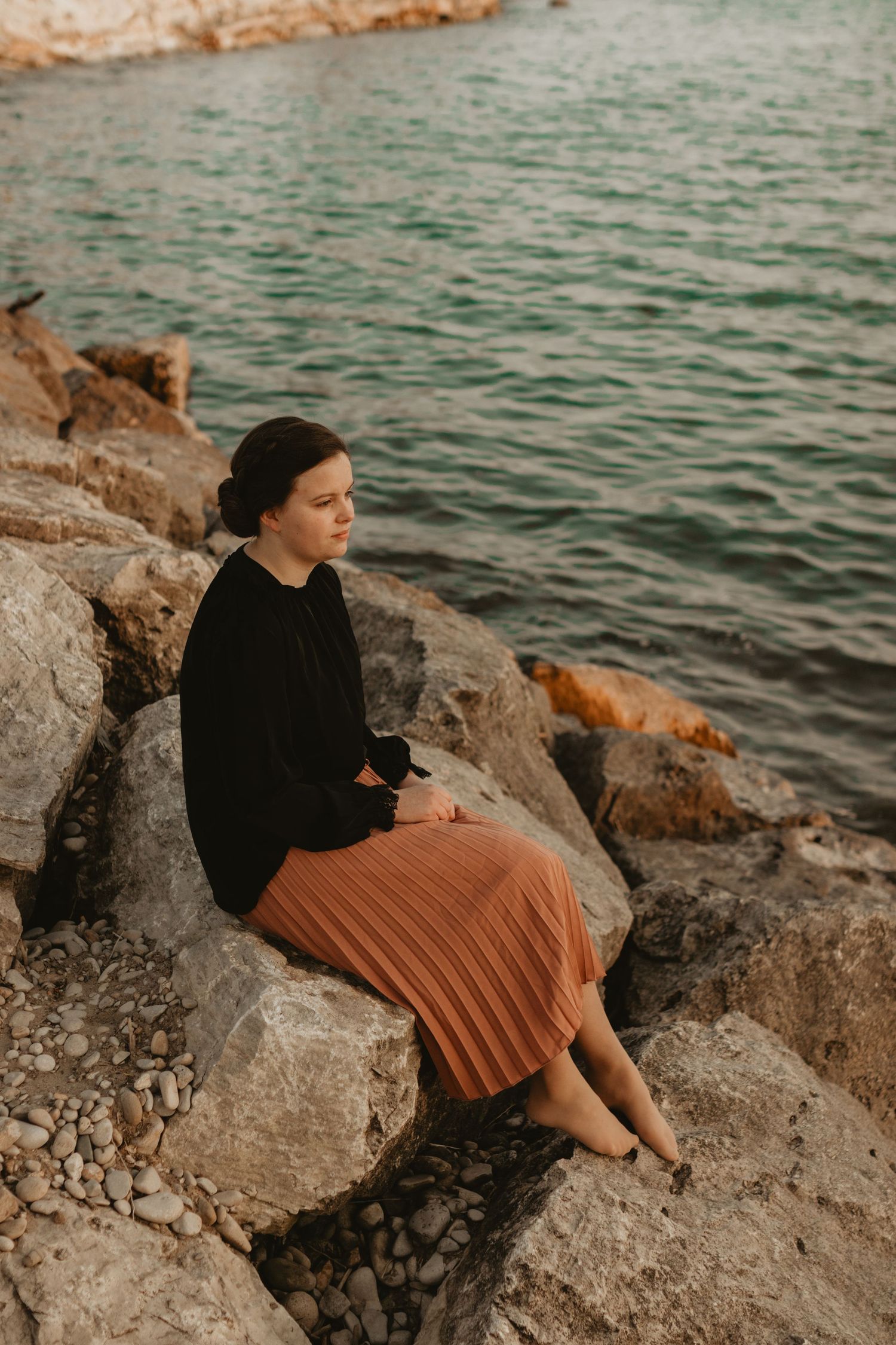 Someone in a black sweater and rust-colored pleated skirt sits contemplatively on coastal rocks by turquoise waters.