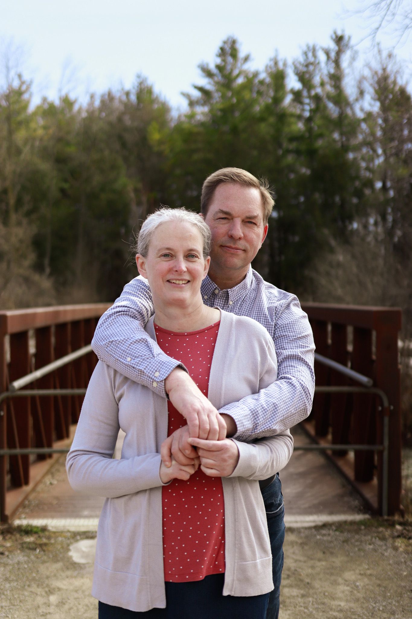 A couple embraces while standing on a wooden bridge with evergreen trees in the background.