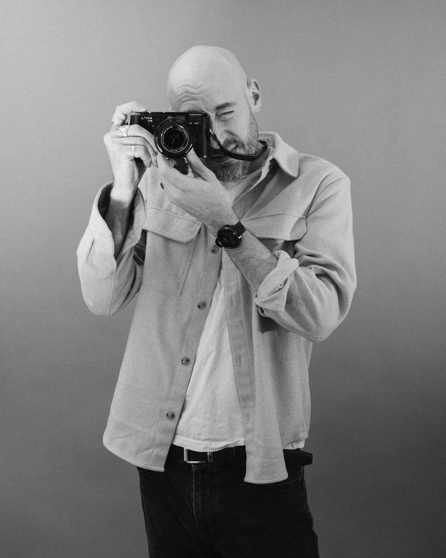 Black and white photo of photographer wearing light button down shirt taking picture with vintage camera against plain background.